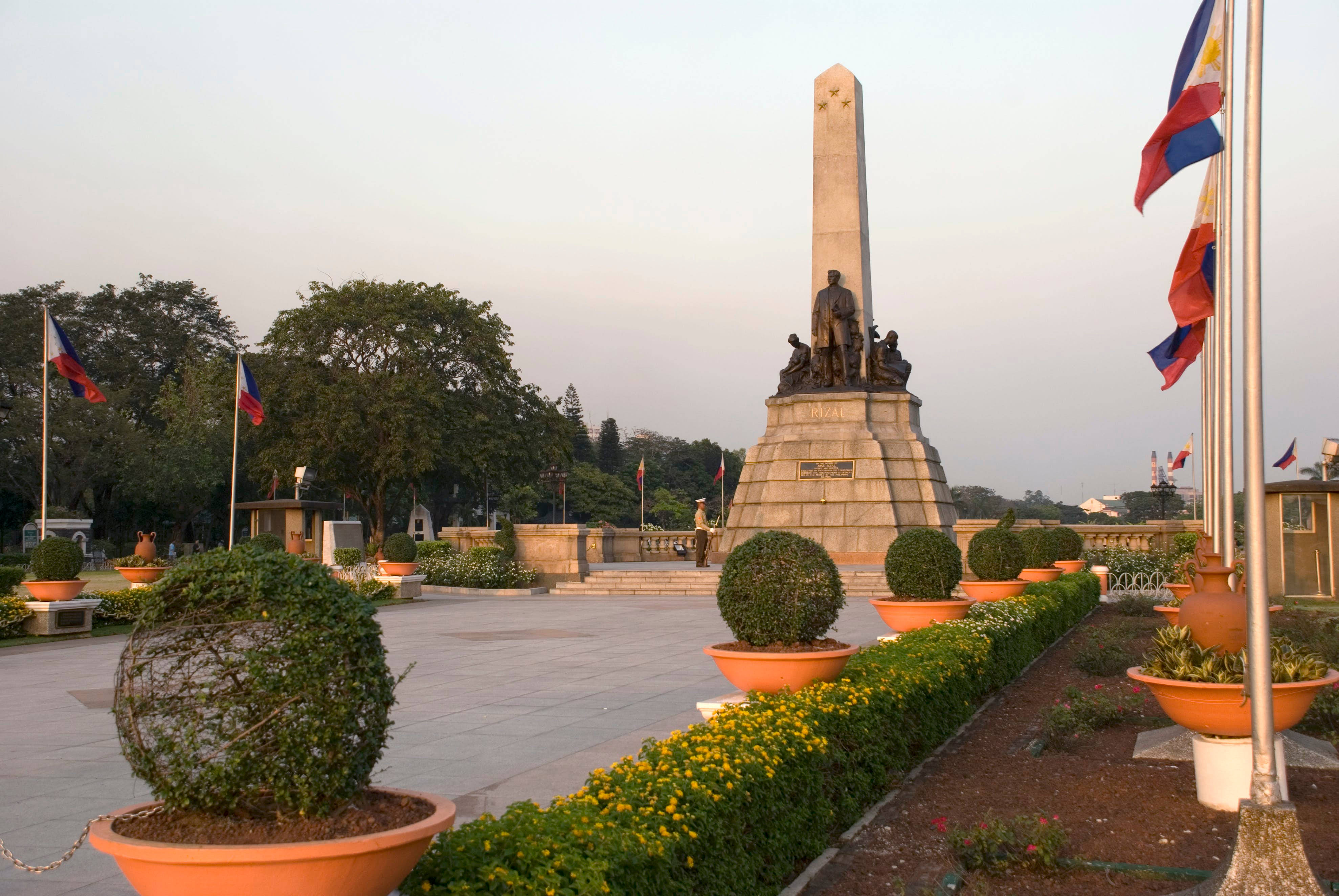 Manila 桌布 壁紙 — 黃昏時分，一個高大的石頭紀念碑，上面有銅像，矗立在鋪砌的廣場上，兩旁擺放著橙色花盆和飄揚的菲律賓國旗。