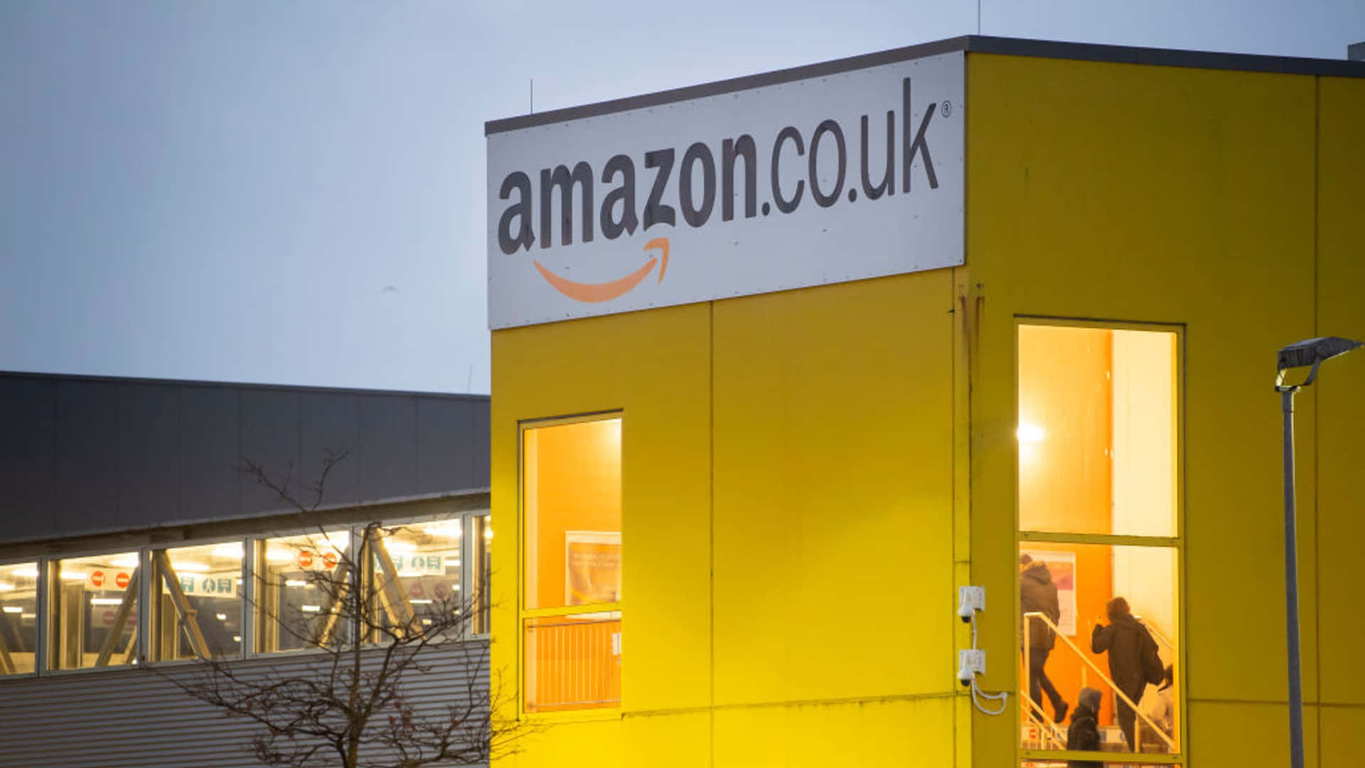 黃昏時分的Amazon UK倉庫外觀，窗戶透出燈光，人們正在進入。 — 亞馬遜 GIF 壁紙