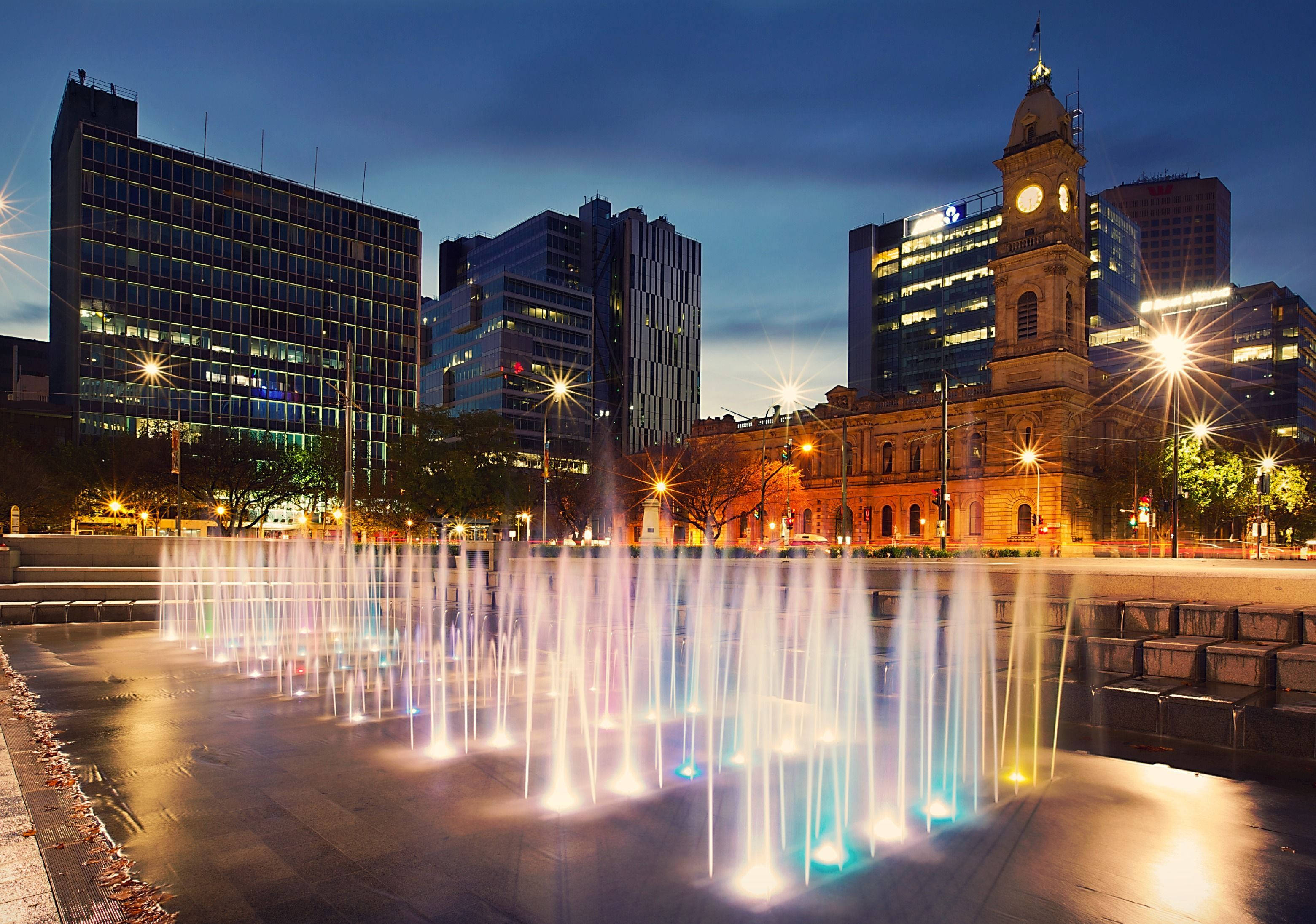 Adelaide 桌布 壁紙 — 黃昏時分，一座充滿活力的城市噴泉在燈火通明的摩天大樓和歷史悠久的鐘樓背景下噴射出色彩繽紛的水柱。