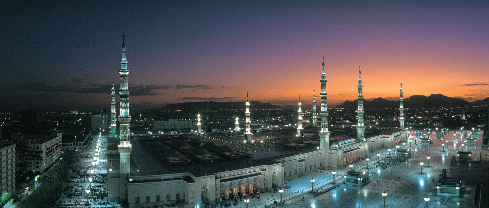 Madina 桌布 壁紙 — 黃昏時分，一座宏偉的清真寺燈火通明，發光的宣禮塔和下方的人群映襯著紫橙色的天空的鳥瞰圖。