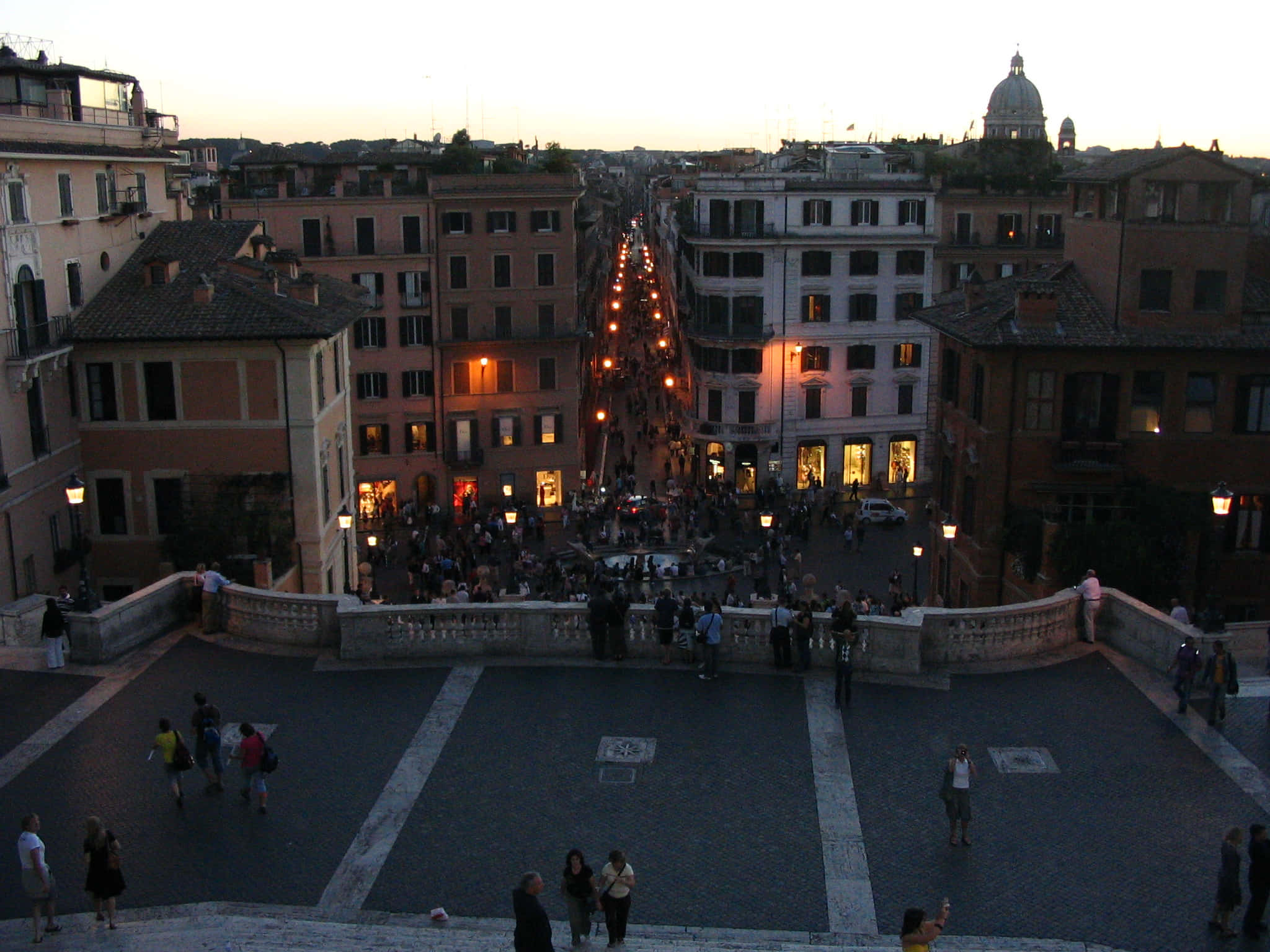 Rome 桌布 壁紙 — 黃昏時分，一條熙來攘往的羅馬街道，窗戶透出溫暖燈光，人們在歷史悠久的石階上漫步。