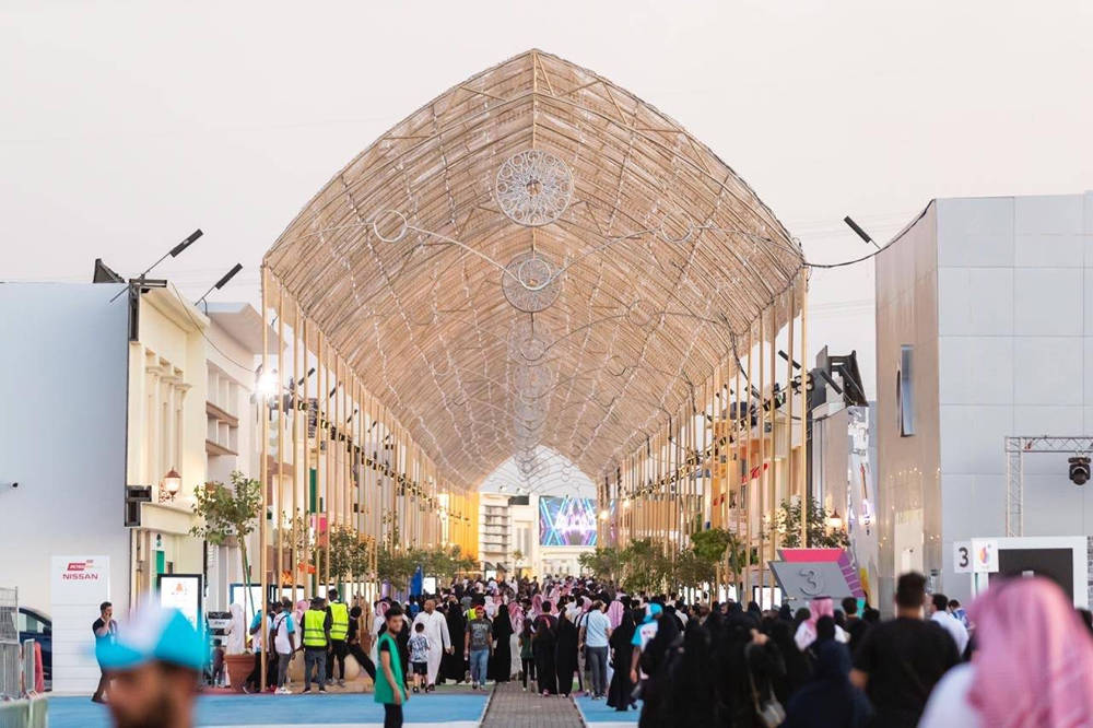 Riyadh 桌布 壁紙 — 黃昏時分，一條熙熙攘攘的行人專用街道，上方是發光的幾何形狀天篷，人群在現代建築之間穿梭。