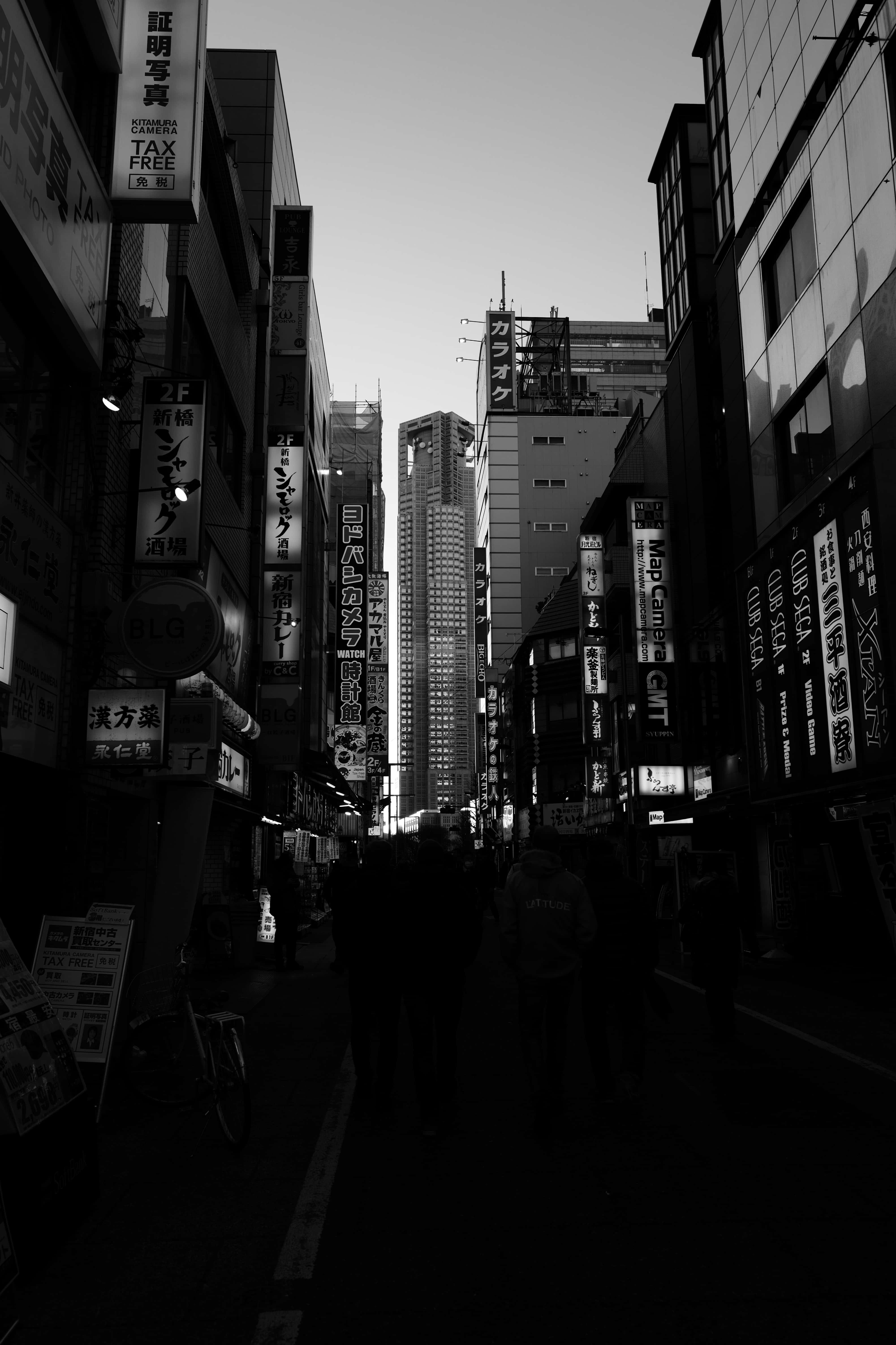 日本黑白 桌布 壁紙 — 黃昏時分，一條狹窄的東京小巷的黑白鮮明景象，高聳的建築物和發光的霓虹燈招牌在剪影般的行人身上投下陰影。