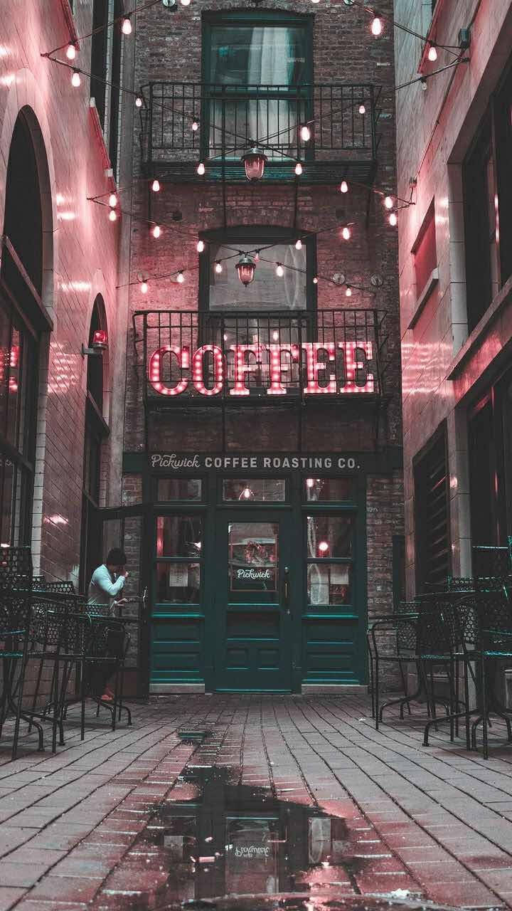 Coffee Shop 桌布 壁紙 — 黃昏時分，一條狹窄的磚砌小巷中，在青綠色門上方有一個發光的粉紅色霓虹燈咖啡店招牌，設有戶外座位和串燈，倒映在水坑中。
