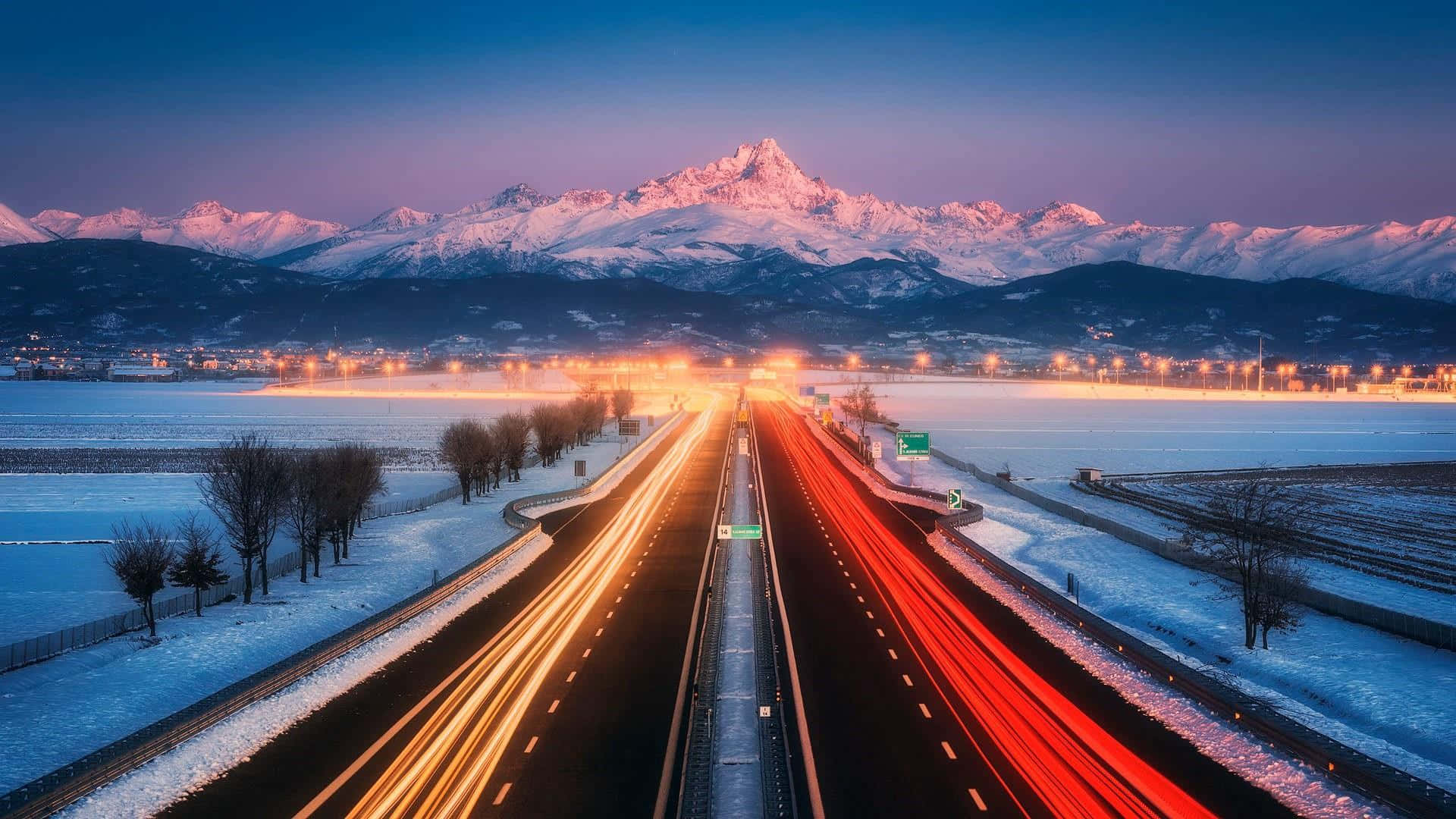 highway 桌布 壁紙 — 黃昏時分，一條積雪的高速公路上，車燈劃出光跡，通往紫色天空下白雪覆蓋的山脈。