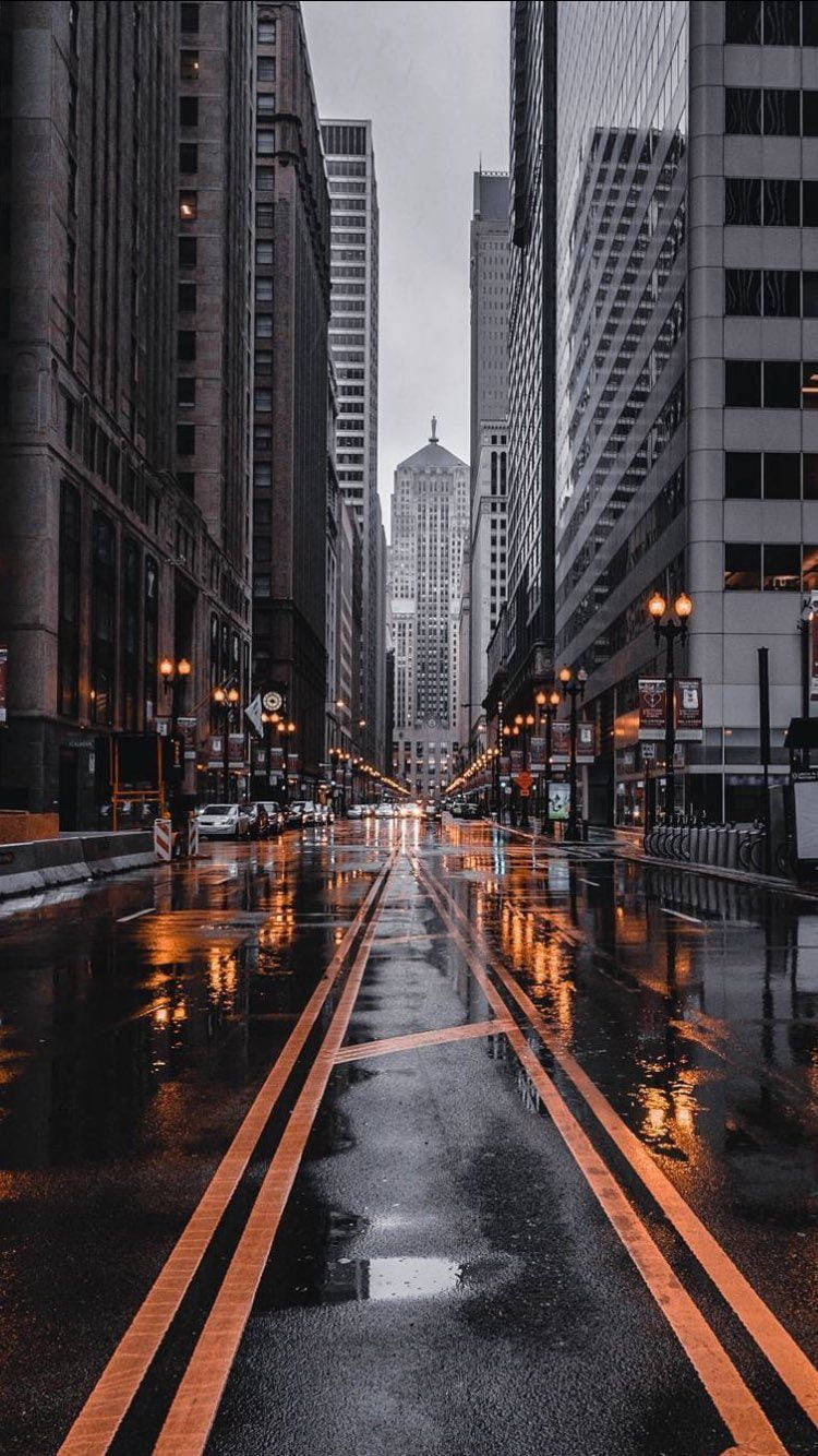 Buildings 桌布 壁紙 — 黃昏時分，一條被雨水打濕的城市街道，溫暖的路燈燈光在高聳的摩天大樓之間的濕漉漉的路面上反射。