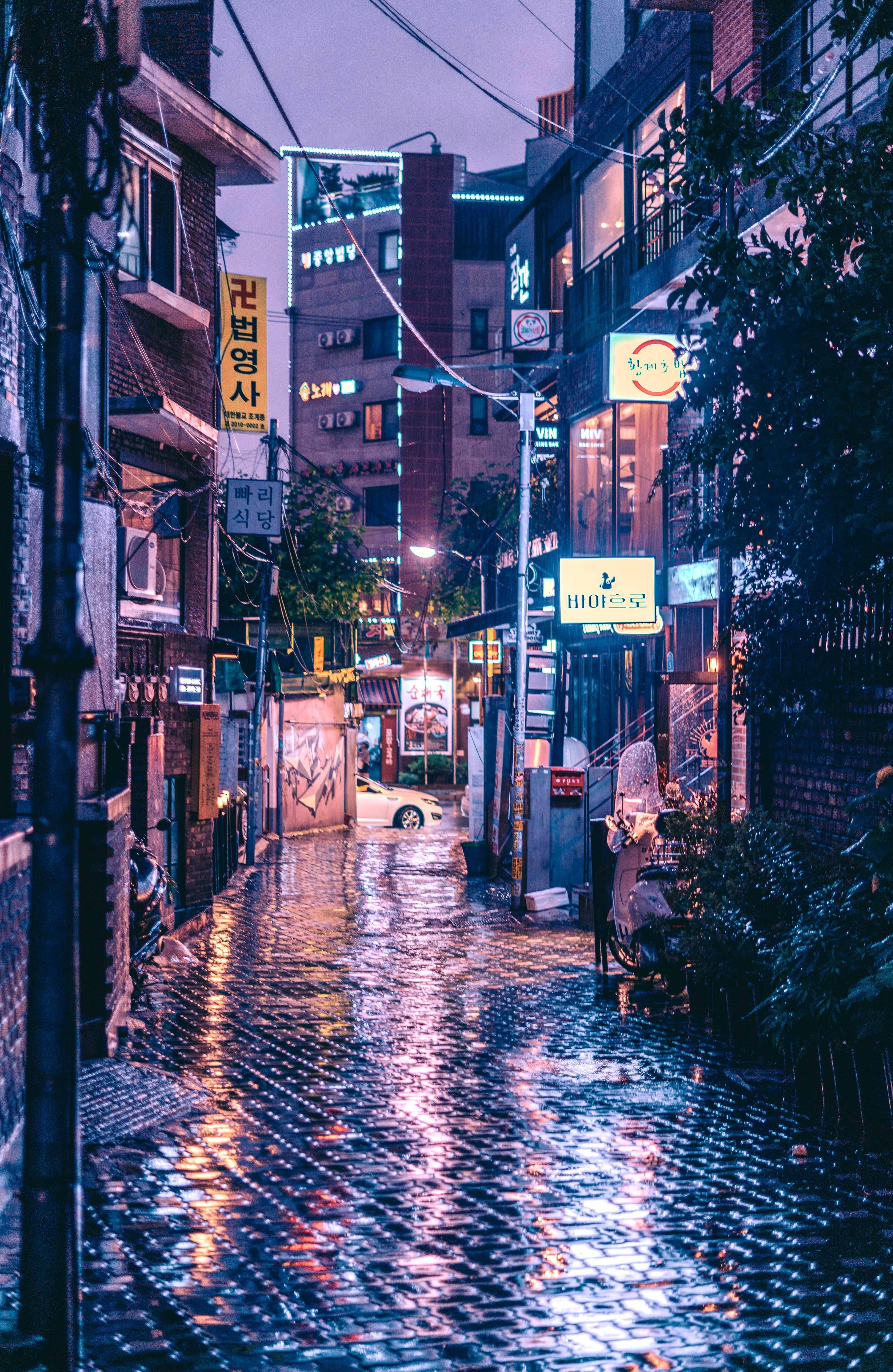 korean 桌布 壁紙 — 黃昏時分，一條被雨水浸濕的城市小巷，在光滑的鵝卵石路上反射出霓虹燈招牌和街燈。