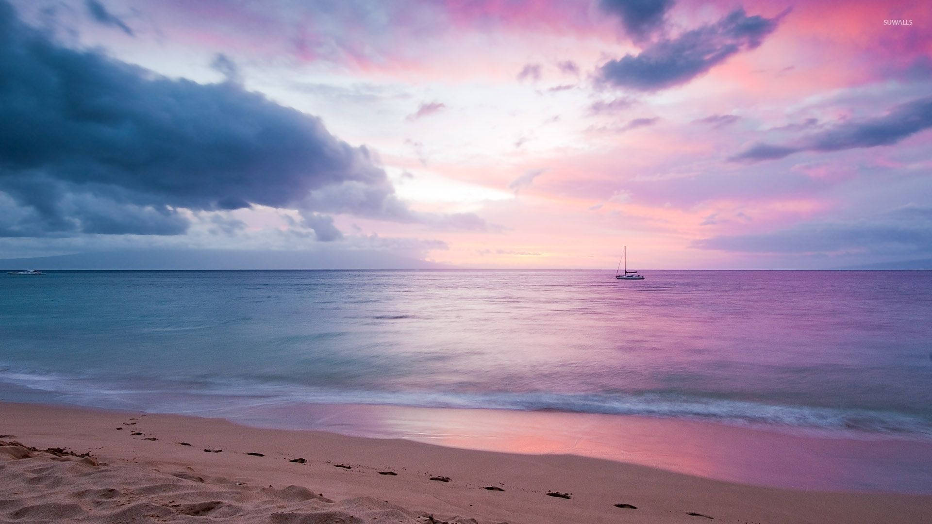 Sunset Aesthetic 桌布 壁紙 — 黃昏時分，一艘孤獨的帆船停泊在平靜的碧綠海面上，粉紅色和紫色的雲彩在帶有腳印的沙灘上空發光。