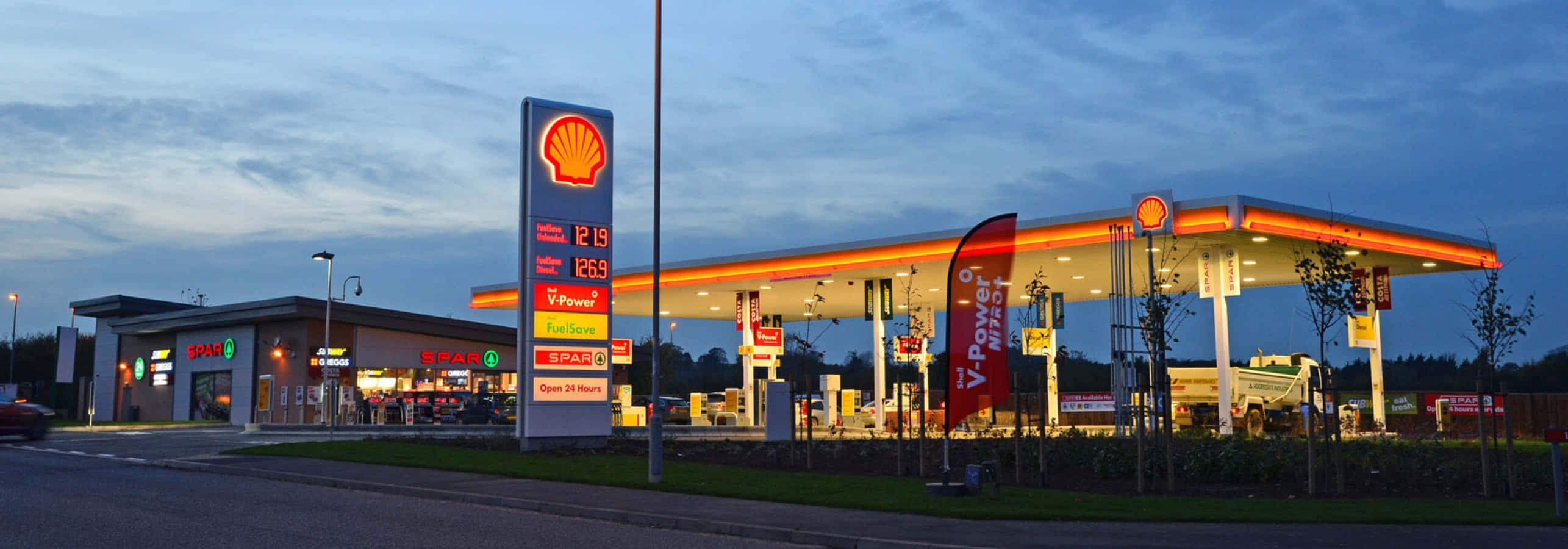 Petrol Station 桌布 壁紙 — 黃昏時分，一間Shell加油站亮起燈光，橙色發光油泵和價格牌映襯著漸暗的藍天。