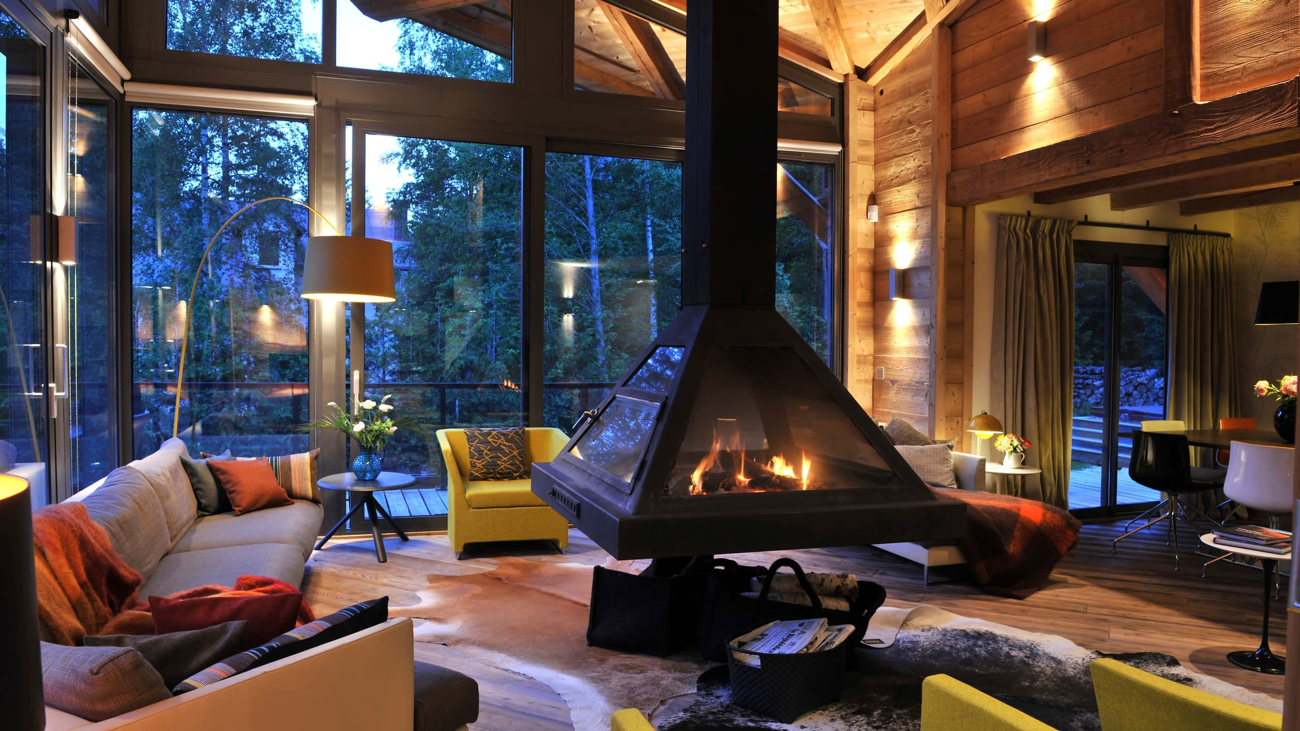 Autumn Fireplace 桌布 壁紙 — 黃昏時分，一間舒適質樸的小木屋客廳，有熊熊燃燒的壁爐和 framing 森林景觀的大窗戶。