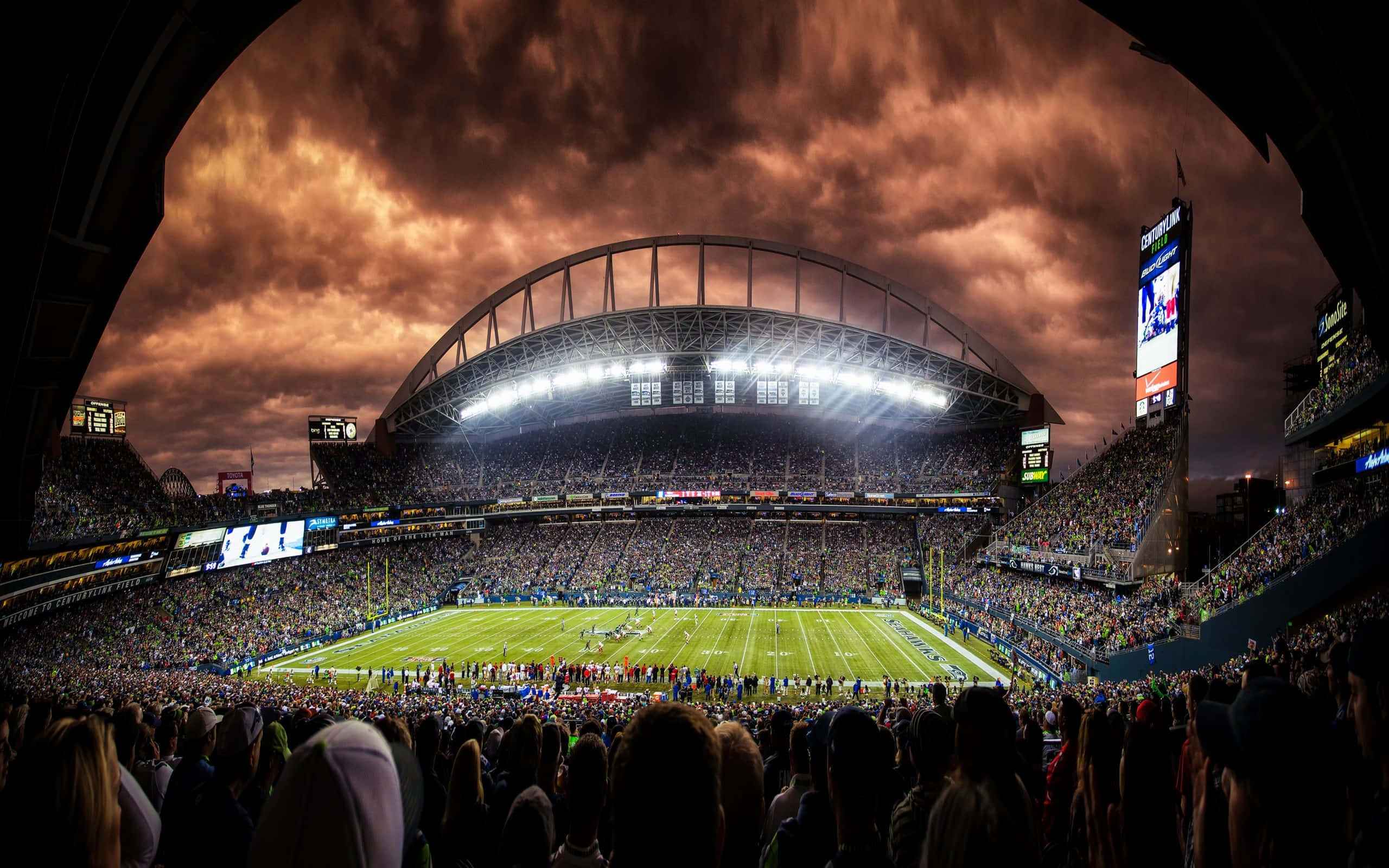 Soccer Stadium 桌布 壁紙 — 黃昏時分，在火紅橙色和暗雲天空下，一個擠滿人的足球場的戲劇性魚眼視角。