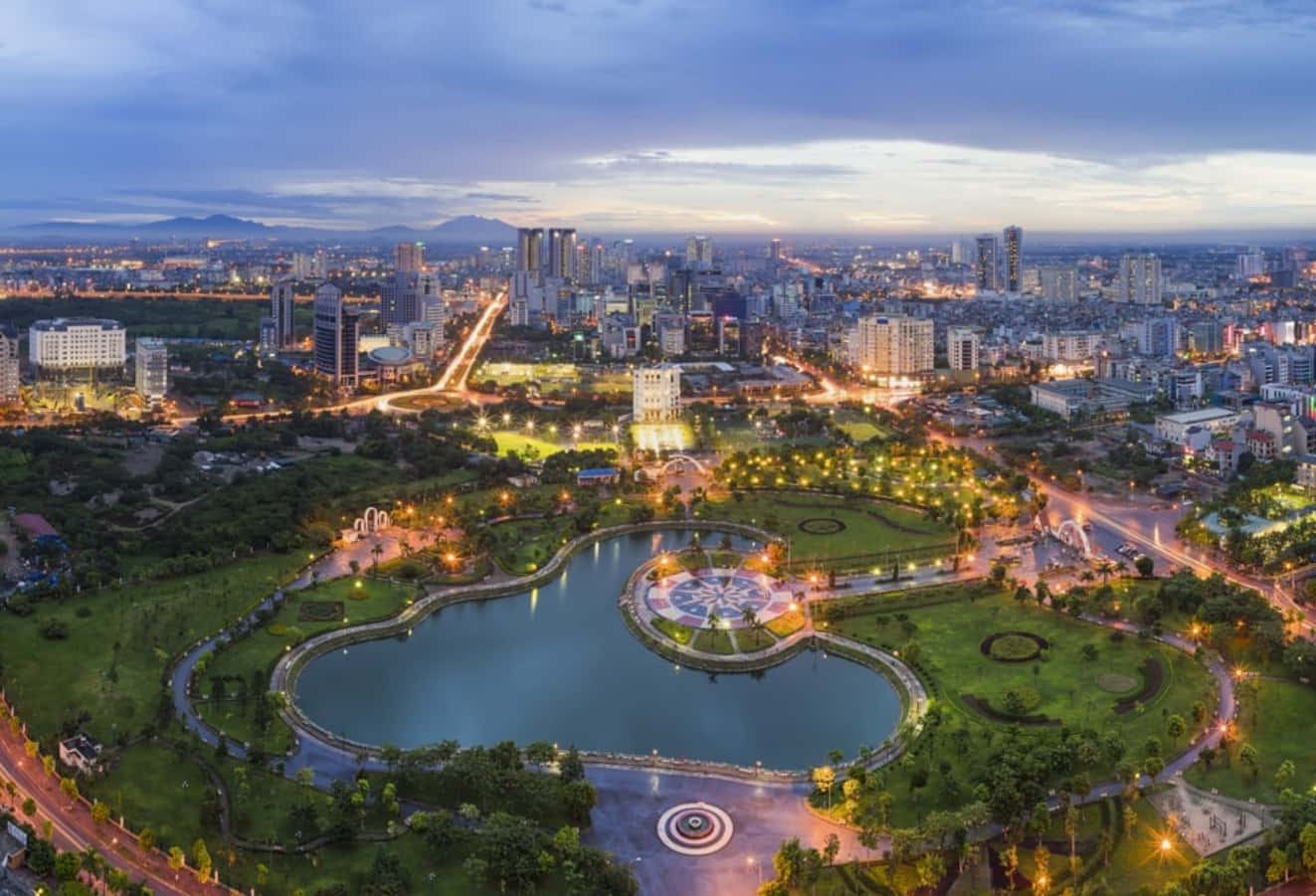 越南 桌布 壁紙 — 黃昏時分，城市公園的鳥瞰圖，有一個心形湖泊，周圍環繞著閃爍的燈光和現代建築，在暮色天空下。