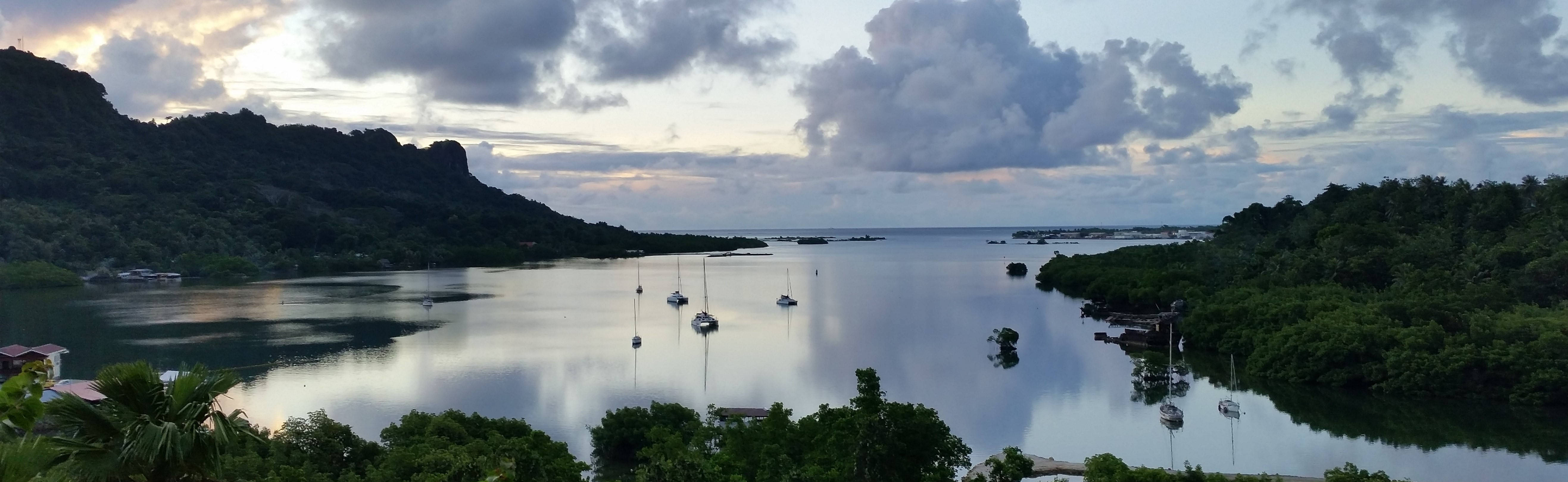 Micronesia 桌布 壁紙 — 黃昏時分，多雲的天空下，鬱鬱蔥蔥的綠色山丘和紅樹林之間停泊著帆船的寧靜熱帶海灣。