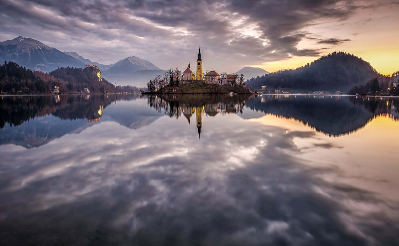 Lake Bled 桌布 壁紙 — 黃昏時分，寧靜的湖面倒映著一座島上的教堂，島嶼被雲層下森林覆蓋的山脈環繞。