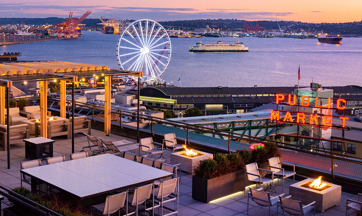 Pike Place Market 桌布 壁紙 — 黃昏時分，屋頂酒廊俯瞰西雅圖海濱，發光的火坑和標誌性的摩天輪在紫色天空的映襯下閃耀。