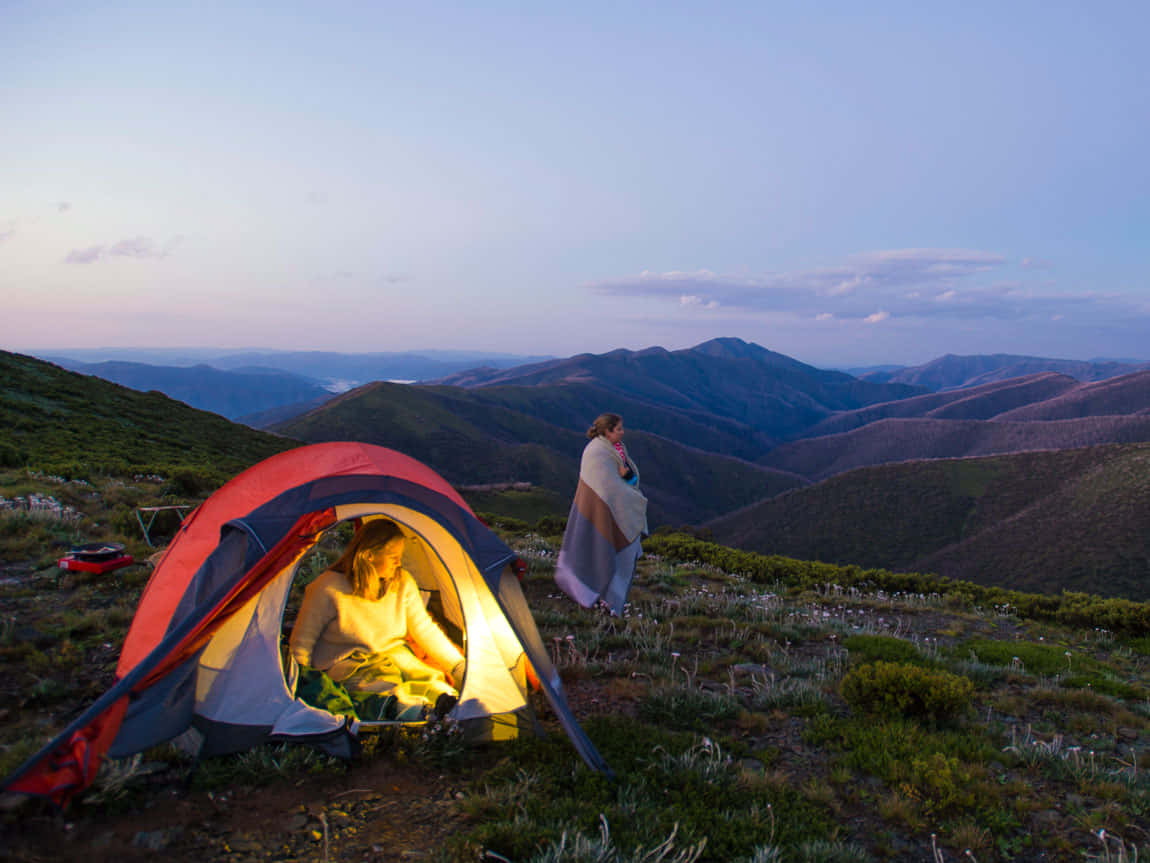 Fall Camping 桌布 壁紙 — 黃昏時分，山脊上一個發光的帳篷，裡面有一個人，附近另一個人裹著毯子。