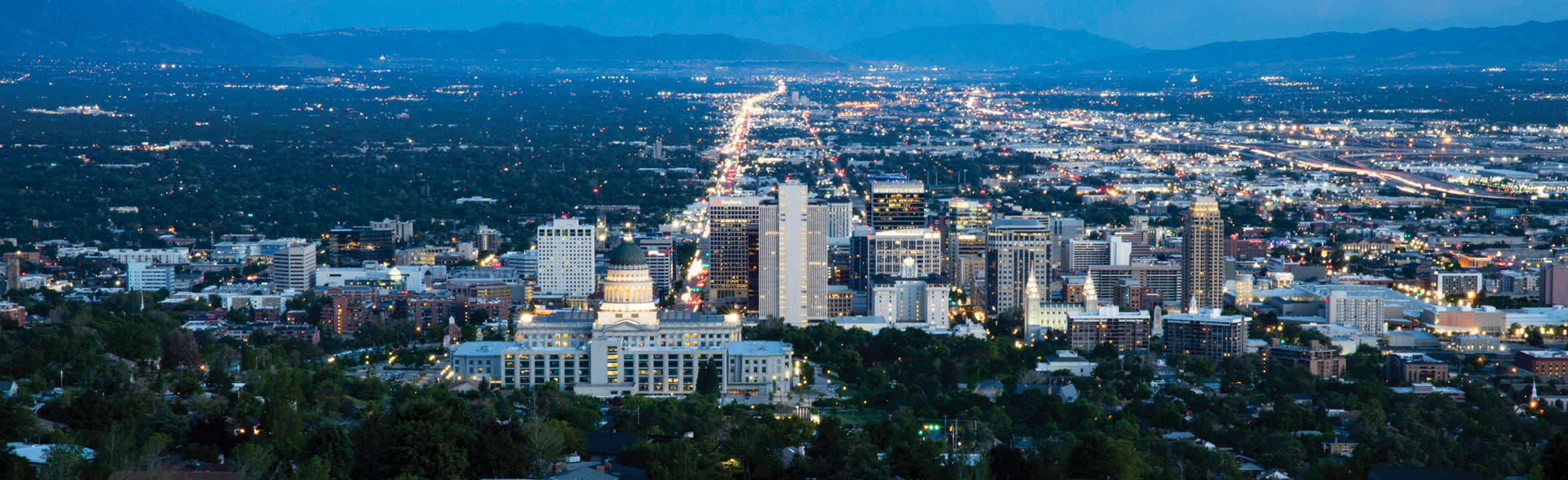Salt Lake City 桌布 壁紙 — 黃昏時分，廣闊的城市景觀，發光的街燈和建築物 nestled 在深綠色的樹木之間，背景有山脈。