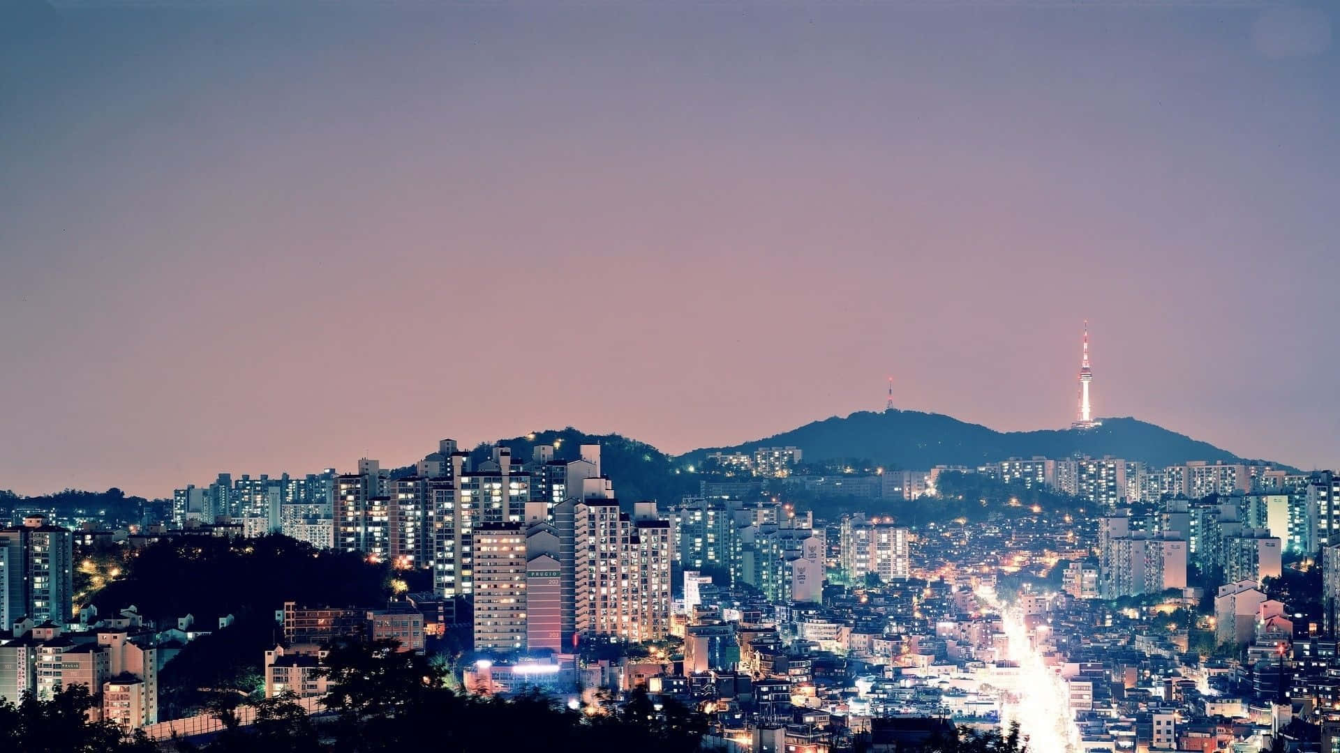 Aesthetic Korean 桌布 壁紙 — 黃昏時分，廣闊的城市景觀，閃爍的摩天大樓和一座高聳的廣播塔在柔和的紫色天空映襯下形成剪影。