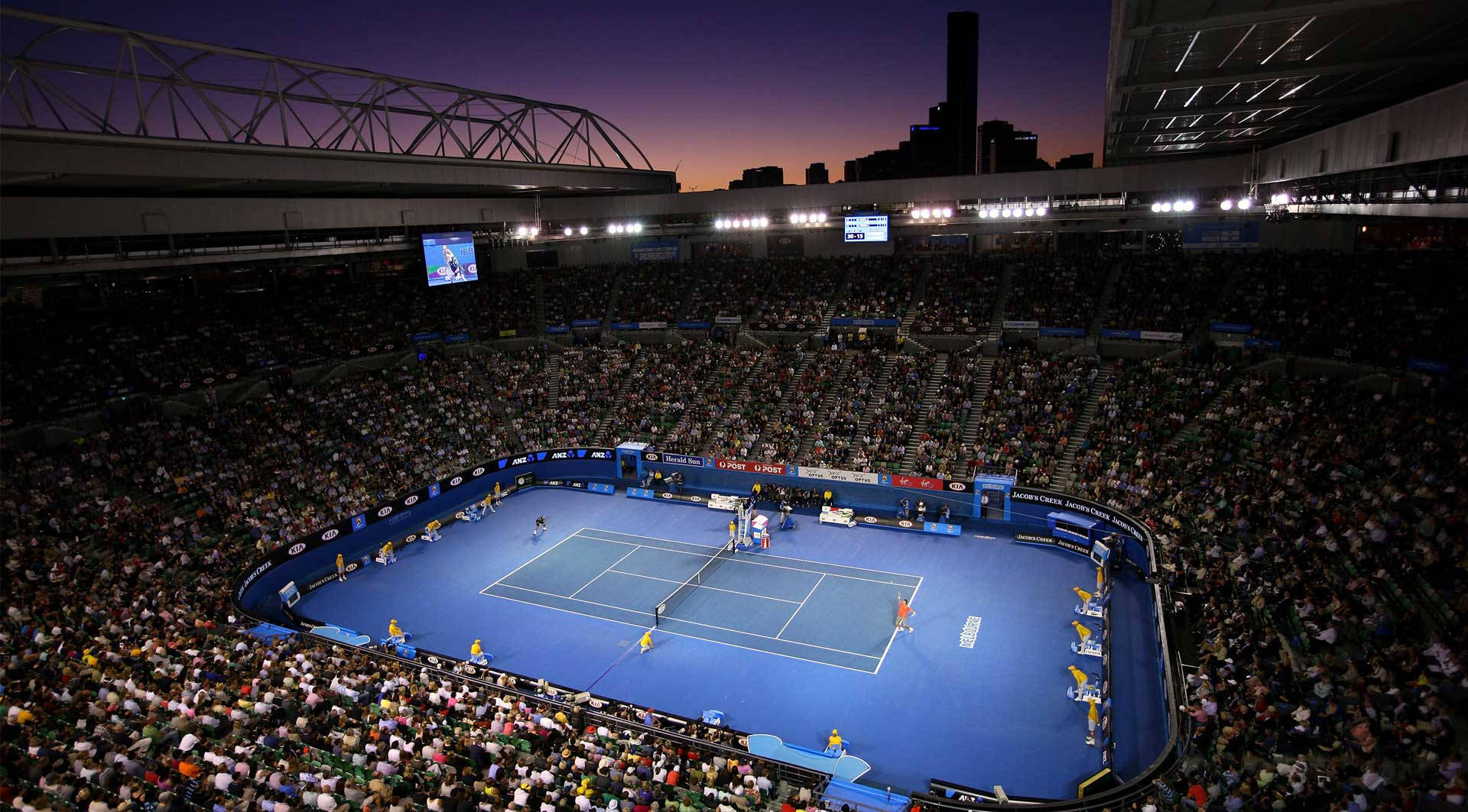 Australian Open 桌布 壁紙 — 黃昏時分，明亮燈光下，藍色球場上球員們在座無虛席的網球場館的鳥瞰圖。