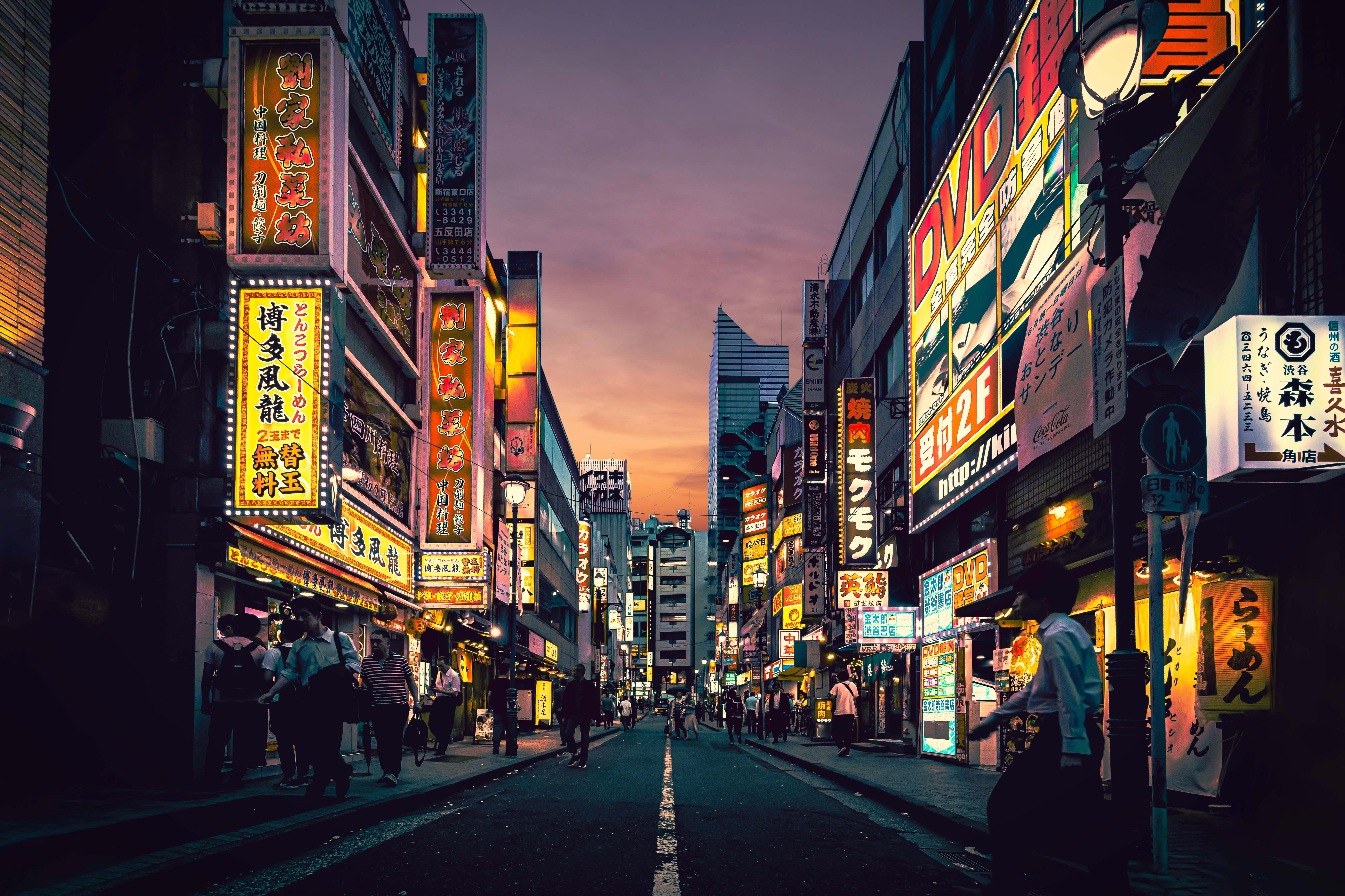 桌布 壁紙 — 黃昏時分，東京一條繁忙的小巷，發光的霓虹燈招牌映照在濕漉漉的人行道上，行人走在高聳的建築物下。