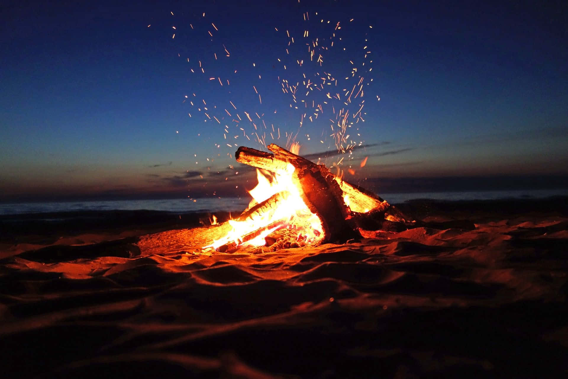 Campfire 桌布 壁紙 — 黃昏時分，沙灘上熊熊燃燒的營火，火花飛向深藍色的天空。