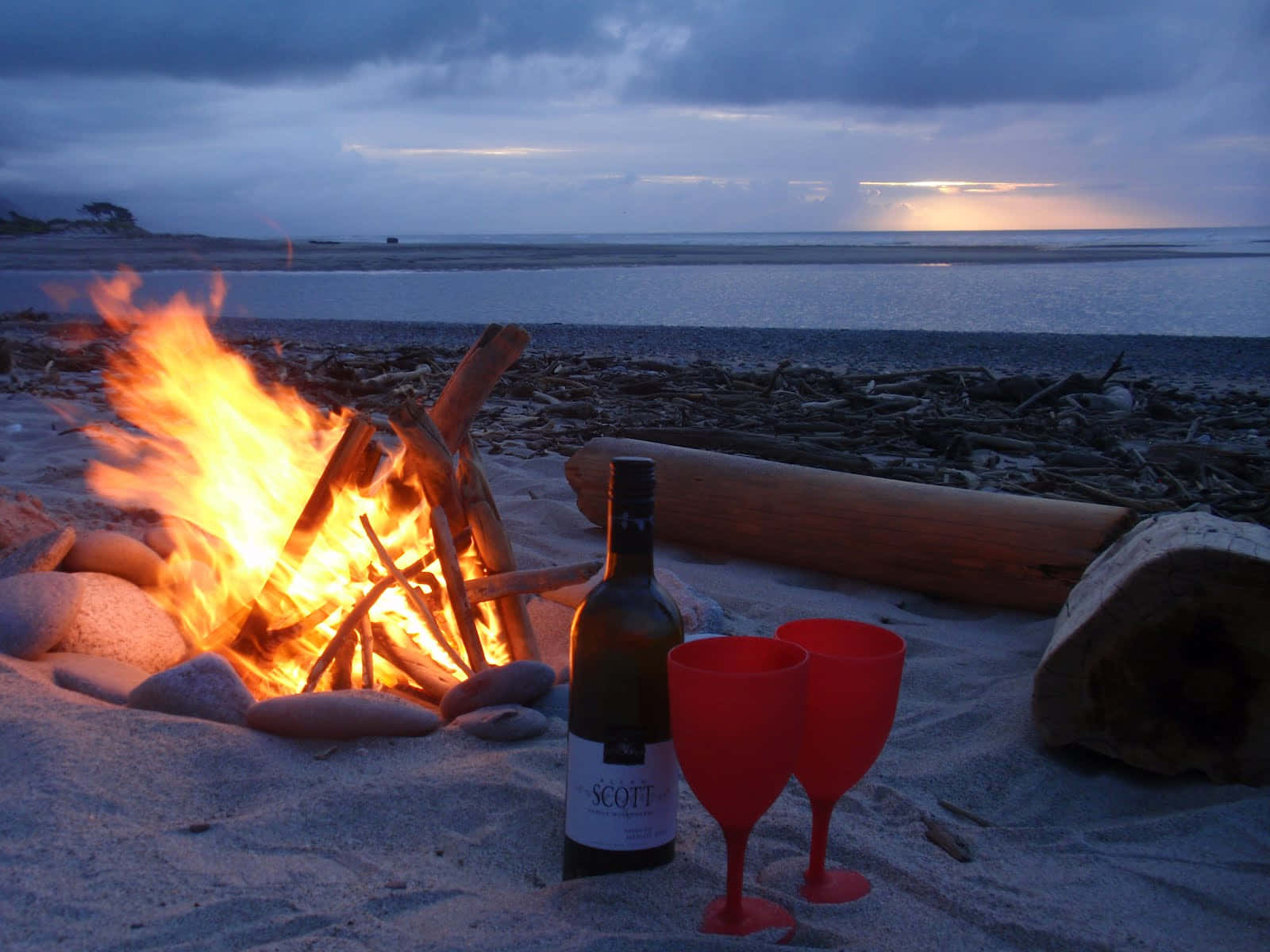 Campfire 桌布 壁紙 — 黃昏時分，沙灘上熊熊燃燒的營火，旁邊有兩杯紅酒杯和一瓶酒，太陽在海洋上落下。
