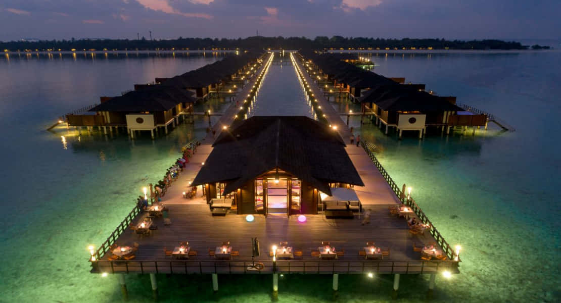 Maldives Island 桌布 壁紙 — 黃昏時分，溫暖燈光映照在碧綠海水上的發光水上平房鳥瞰圖。