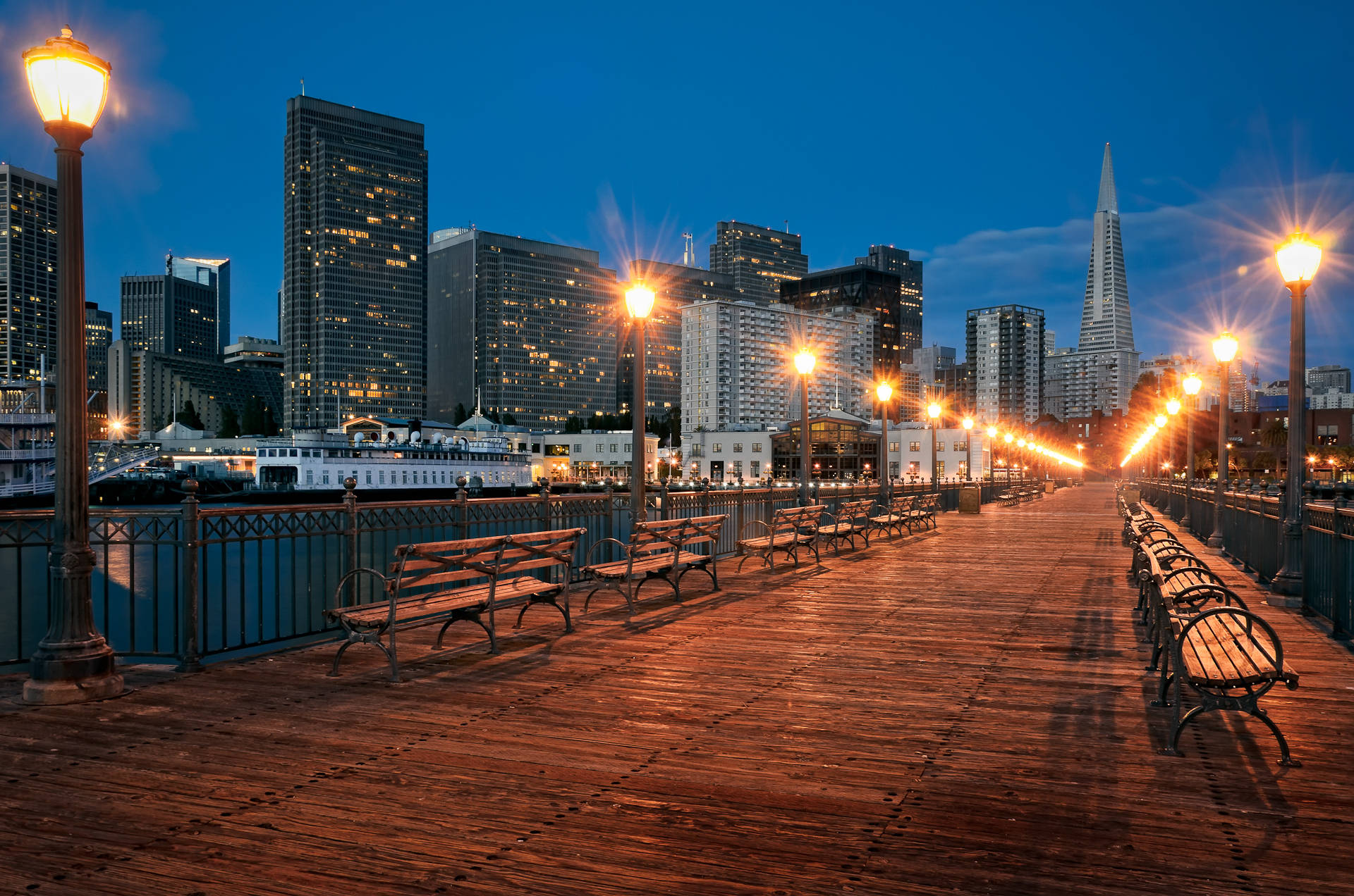 加州 桌布 壁紙 — 黃昏時分，溫暖燈光照亮的木製碼頭，閃爍的路燈，背景是包括泛美金字塔在內的城市天際線。