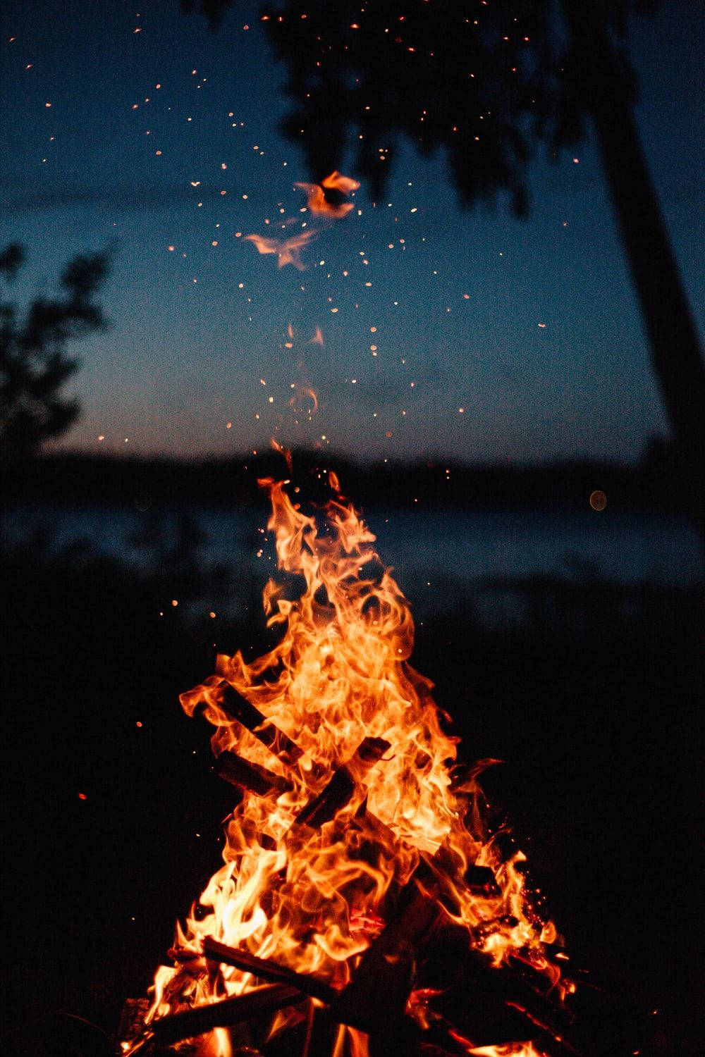 Flame 桌布 壁紙 — 黃昏時分，熊熊營火將火花射向深藍色的暮色天空，樹木剪影清晰可見。