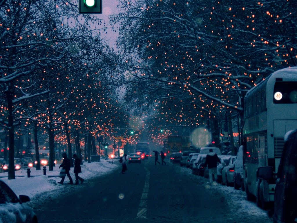 冬季美學 桌布 壁紙 — 黃昏時分，白雪覆蓋城市街道，溫暖的串燈在光禿的樹木間閃爍，行人走在綠色交通燈下。