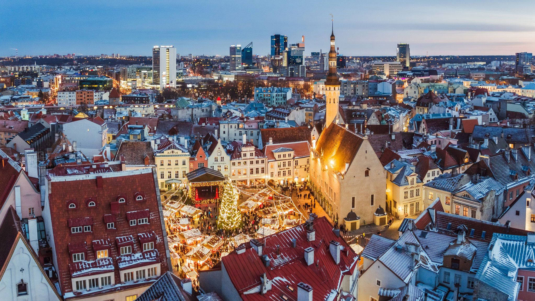 Estonia 桌布 壁紙 — 黃昏時分，白雪覆蓋的歐洲城市廣場鳥瞰圖，閃爍的聖誕市集攤位和歷史建築。