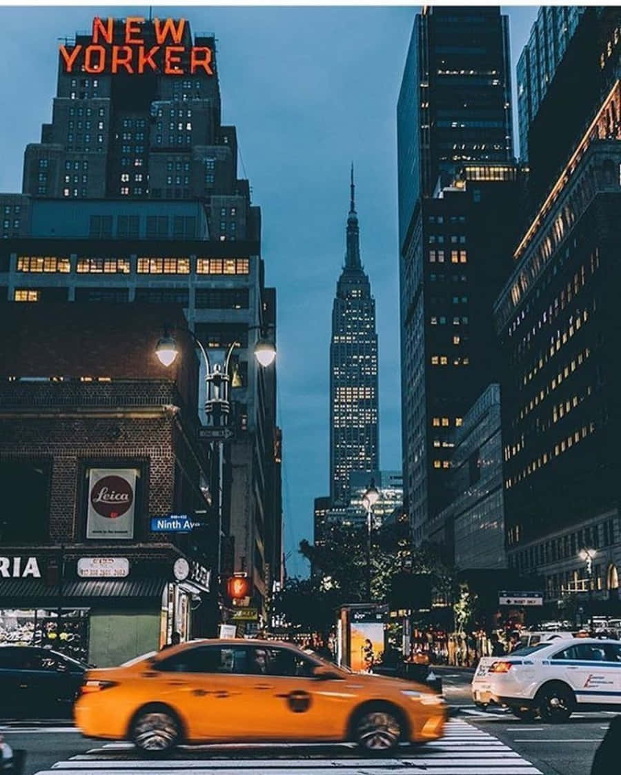 Nyc Aesthetic 桌布 壁紙 — 黃昏時分，紐約市一條充滿活力的街道，一輛黃色計程車飛馳而過高聳的摩天大樓，遠處標誌性的帝國大廈閃閃發光。