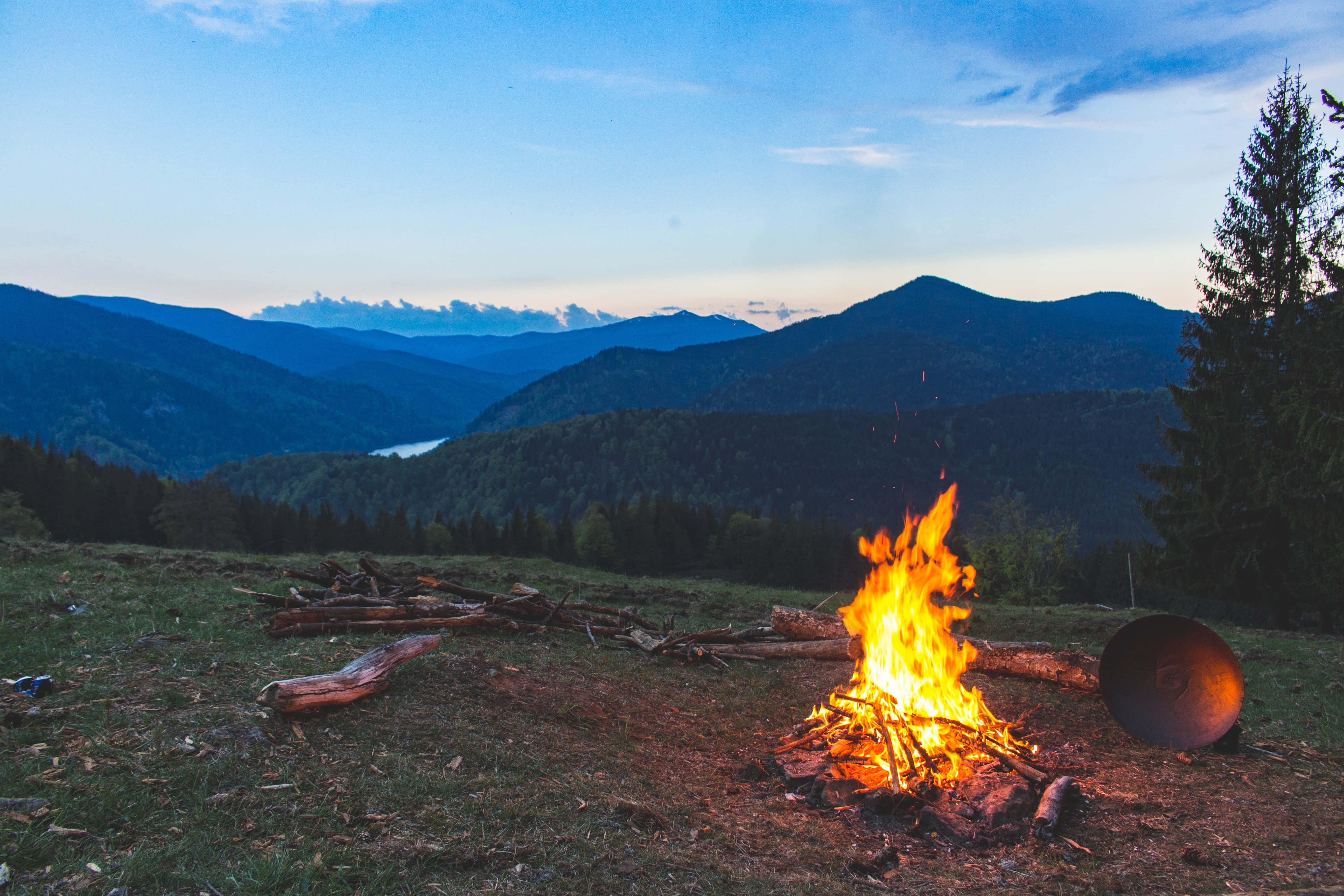 Bonfire 桌布 壁紙 — 黃昏時分，草地上的一個營火在丘陵上噼啪作響，遠方是層層疊疊的山脈和寧靜的湖泊。