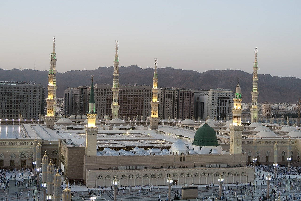 Madina Sharif 桌布 壁紙 — 黃昏時分，被山脈和人群環繞的宏偉清真寺，帶有發光的宣禮塔和綠色圓頂。
