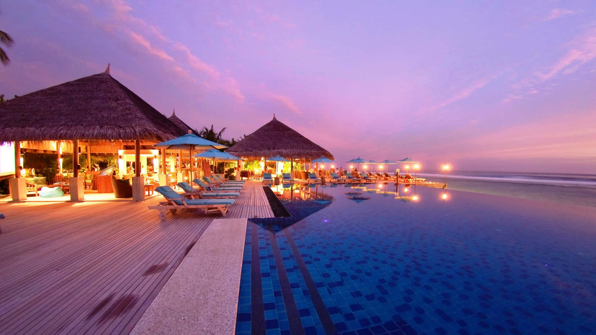 Maldives Beach 桌布 壁紙 — 黃昏時分，豪華的無邊際泳池映照著紫色天空和海洋旁發光的茅草屋。