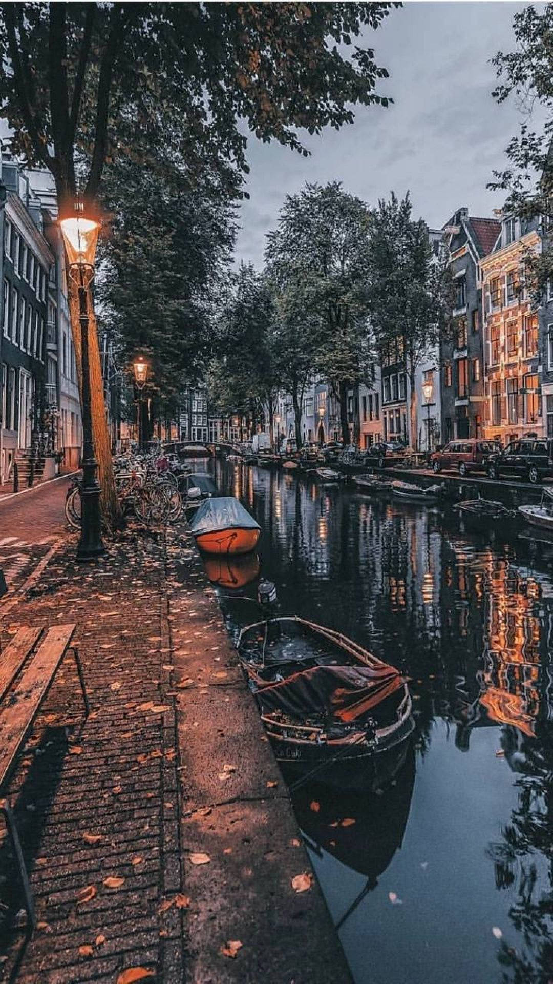 Amsterdam 桌布 壁紙 — 黃昏時分，阿姆斯特丹一條寧靜的運河，發光的街燈倒映在平靜的水面上，旁邊是歷史建築和停泊的船隻。