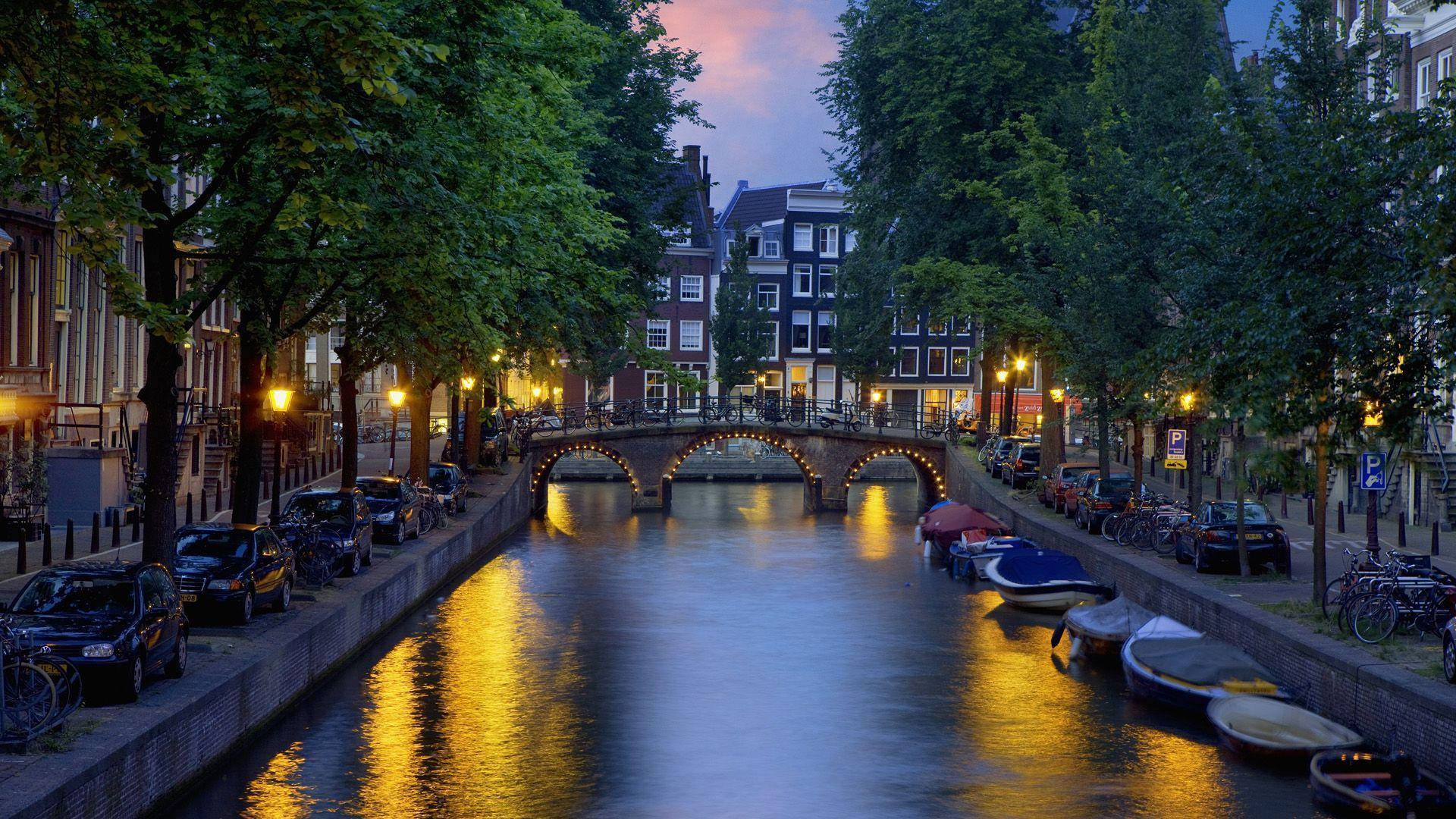 Amsterdam 桌布 壁紙 — 黃昏時分，阿姆斯特丹的運河寧靜祥和，溫暖的街燈倒映在石橋下的平靜水面上。