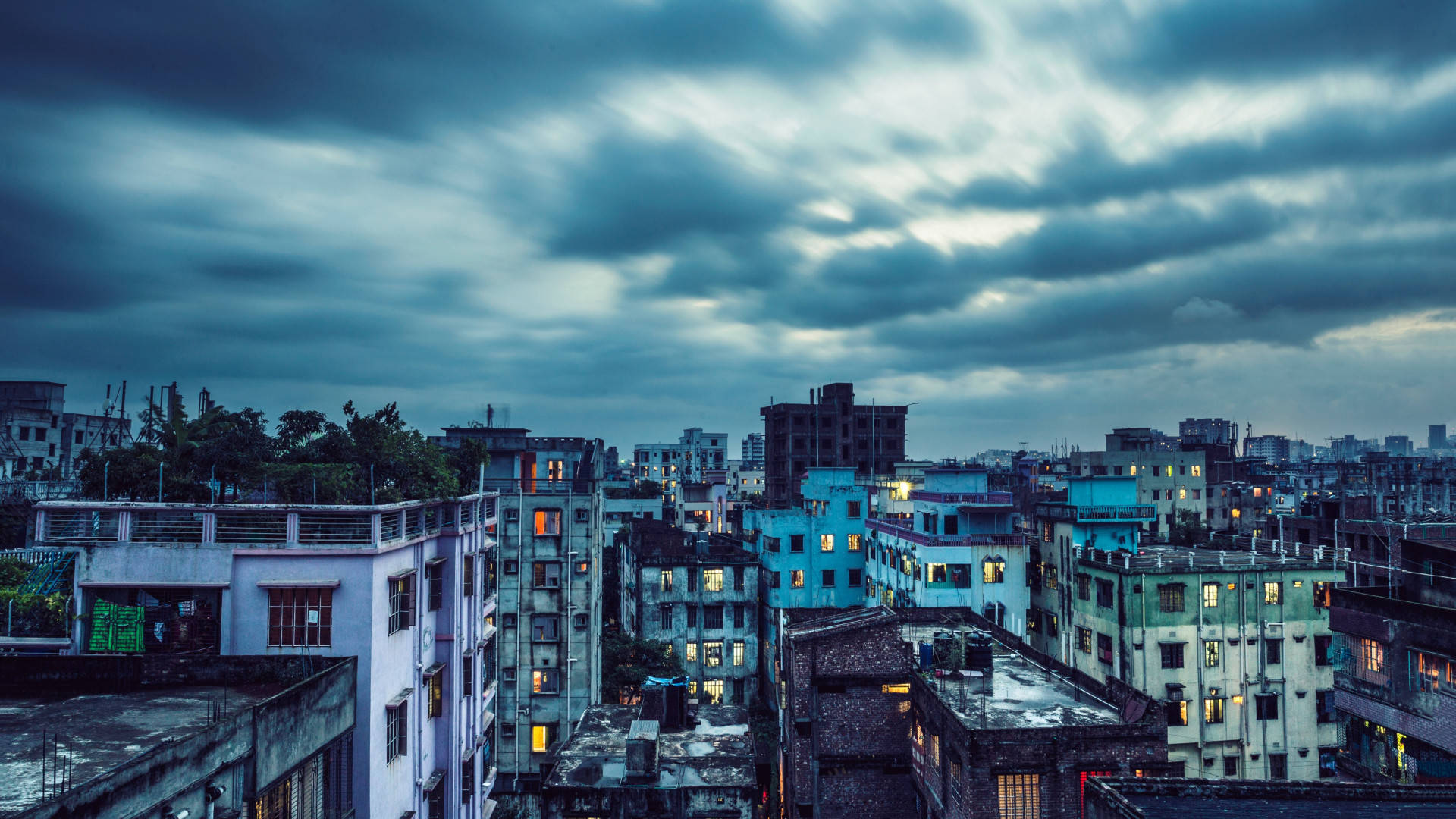 Bangladesh 桌布 壁紙 — 黃昏時分，陰鬱的城市天際線，帶有條紋雲彩，溫暖的燈光從飽經風霜的建築物窗戶中閃爍。