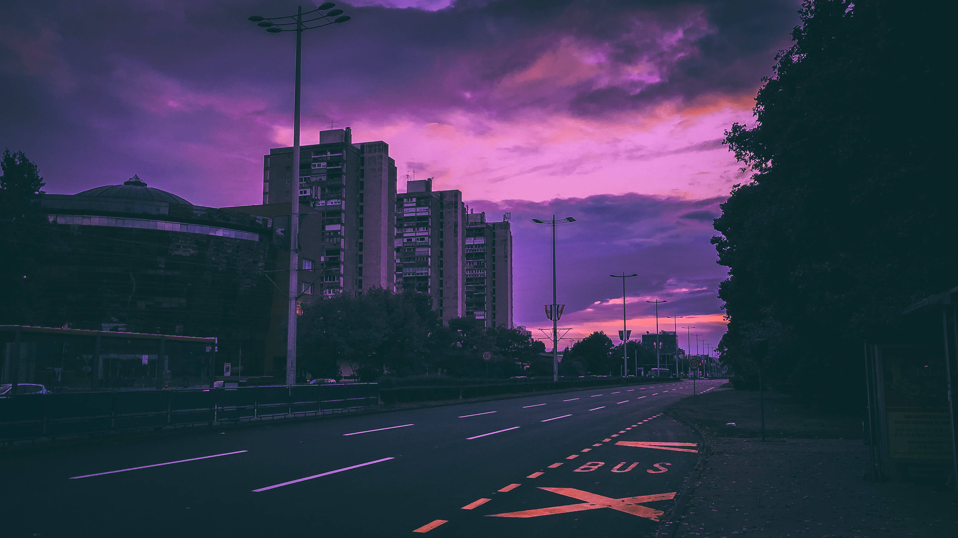 Road 桌布 壁紙 — 黃昏時分一條寧靜的城市街道，沐浴在充滿活力的紫色和粉紅色天空下，高樓大廈在背景中聳立。