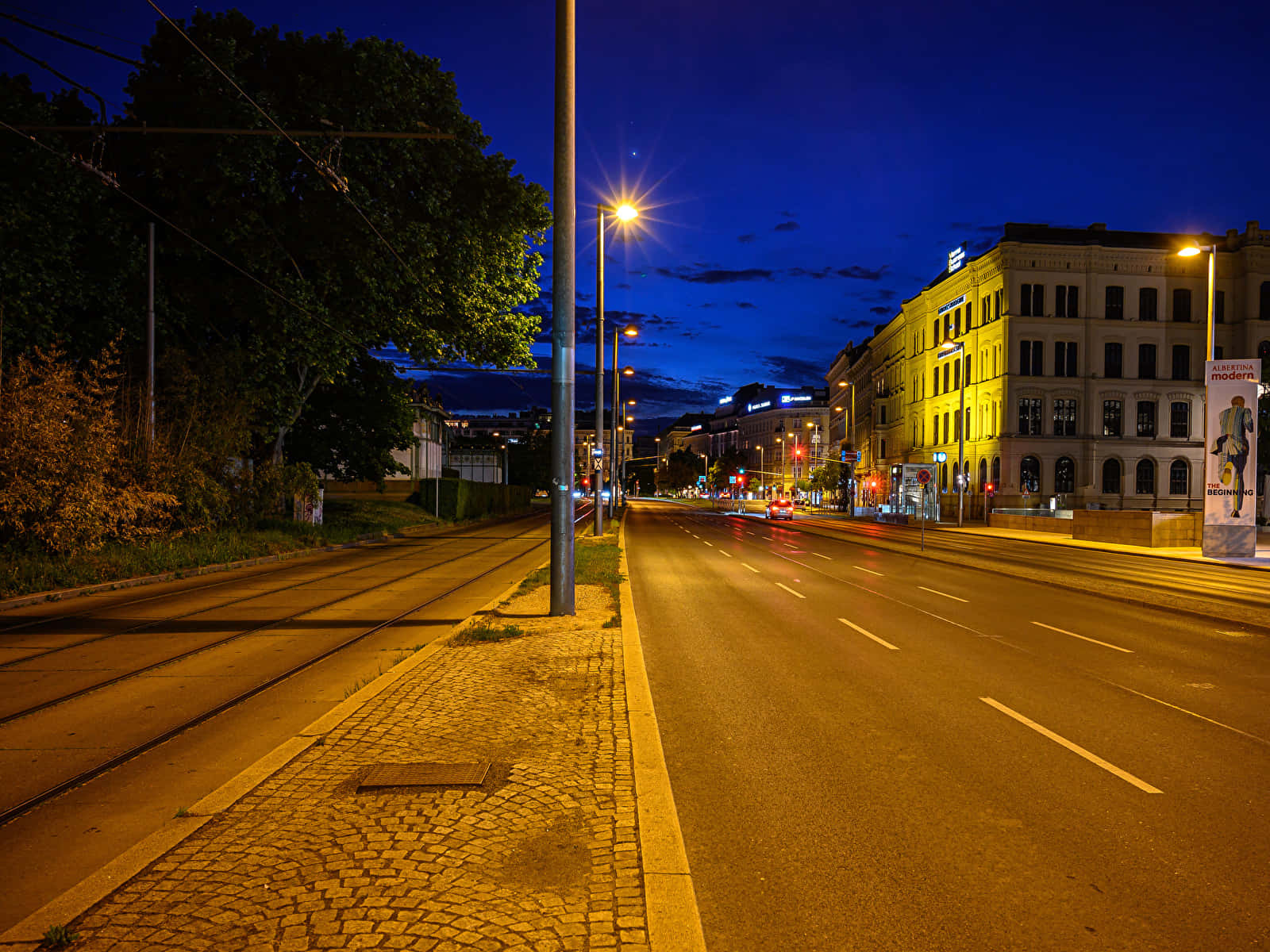 Street Light 桌布 壁紙 — 黃昏時分一條寧靜的城市街道，溫暖的路燈照亮了鵝卵石人行道和電車軌道，天空呈現深藍色。