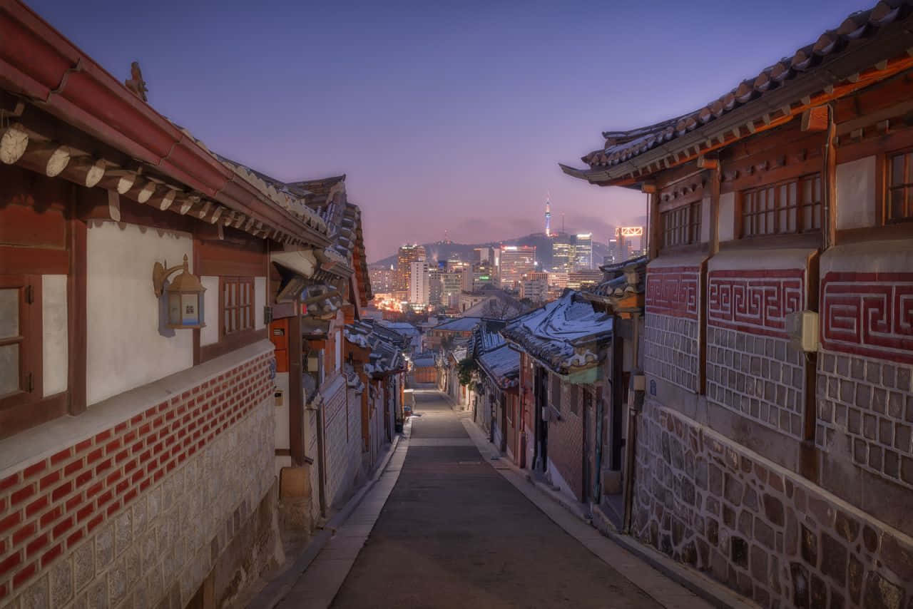 Aesthetic Korean 桌布 壁紙 — 黃昏時分一條狹窄的傳統韓國小巷，屋頂上覆蓋著雪花，遠處是閃爍的現代城市天際線。
