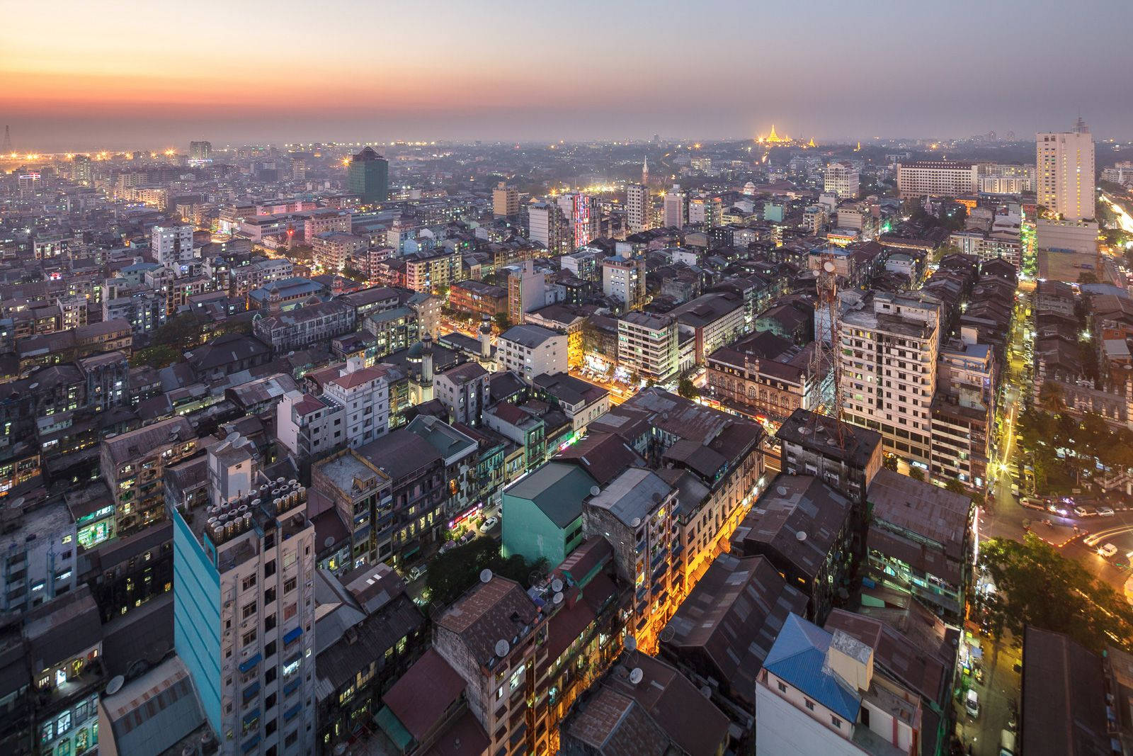 Yangon 桌布 壁紙 — 黃昏時分俯瞰一座廣闊的城市，溫暖的燈光從建築物和街道中閃耀，天空呈現柔和的橙色和紫色。