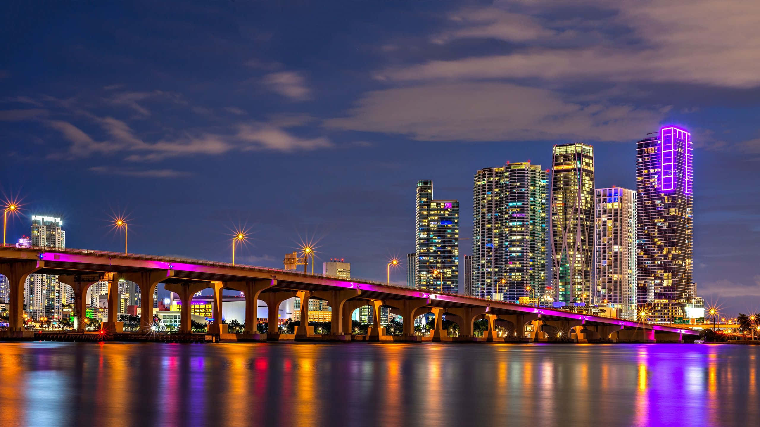 Miami 4K 桌布 壁紙 — 黃昏時分充滿活力的城市天際線，一座發光的橋樑在平靜的水面上反射著霓虹燈光。