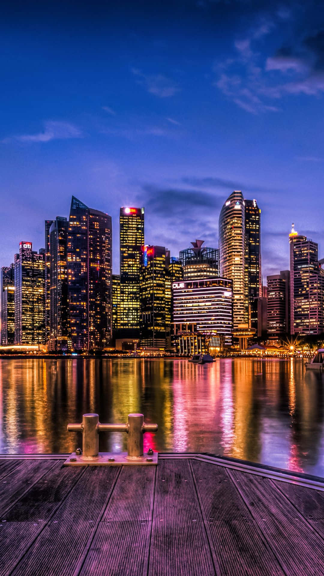 Singapore 桌布 壁紙 — 黃昏時分充滿活力的城市天際線，前景的木製碼頭映照著燈火通明的摩天大樓於平靜的水面上。