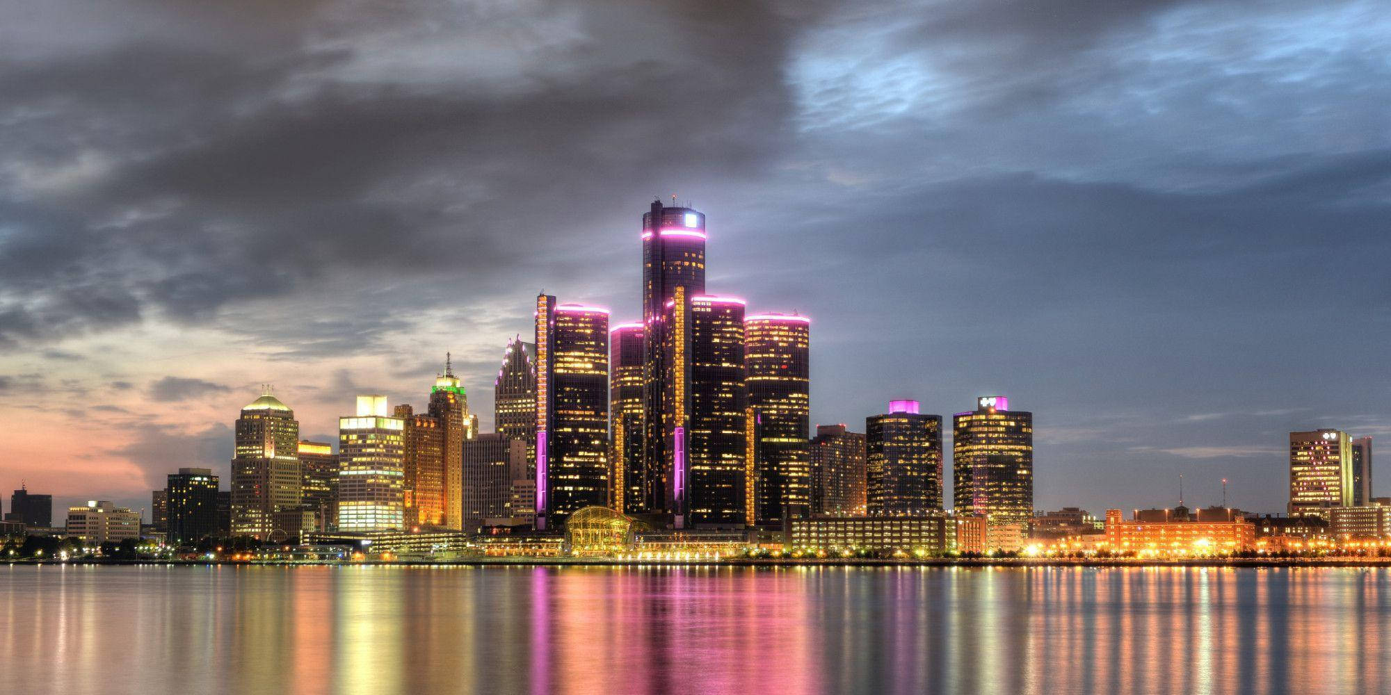 detroit 桌布 壁紙 — 黃昏時分充滿活力的城市天際線，摩天大樓在粉紅色和金色的燈光中閃耀，倒映在平靜的水面上，天空陰沉多雲。
