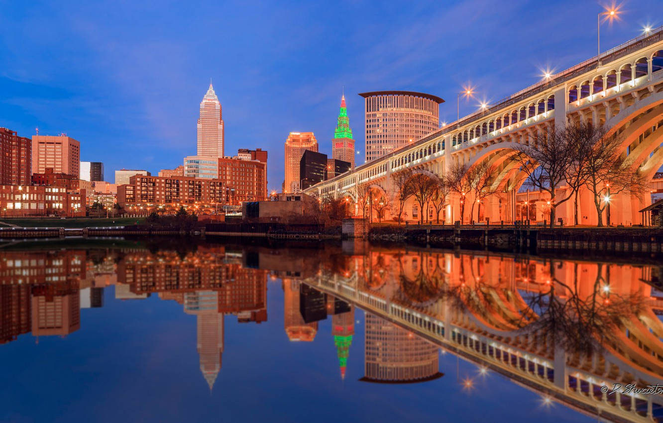 夜空 桌布 壁紙 — 黃昏時分充滿活力的城市天際線，燈火通明的建築物和橋樑倒映在平靜的水面上。
