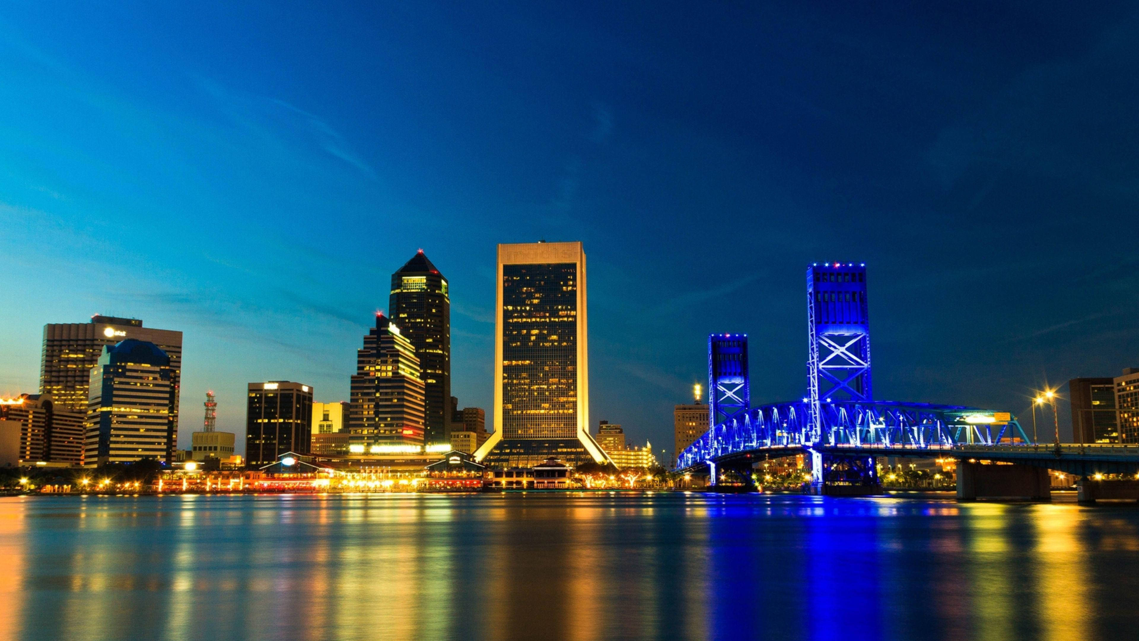 Jacksonville 桌布 壁紙 — 黃昏時分充滿活力的城市天際線，燈火通明的摩天大樓和發光的藍色橋樑倒映在平靜的水面上。