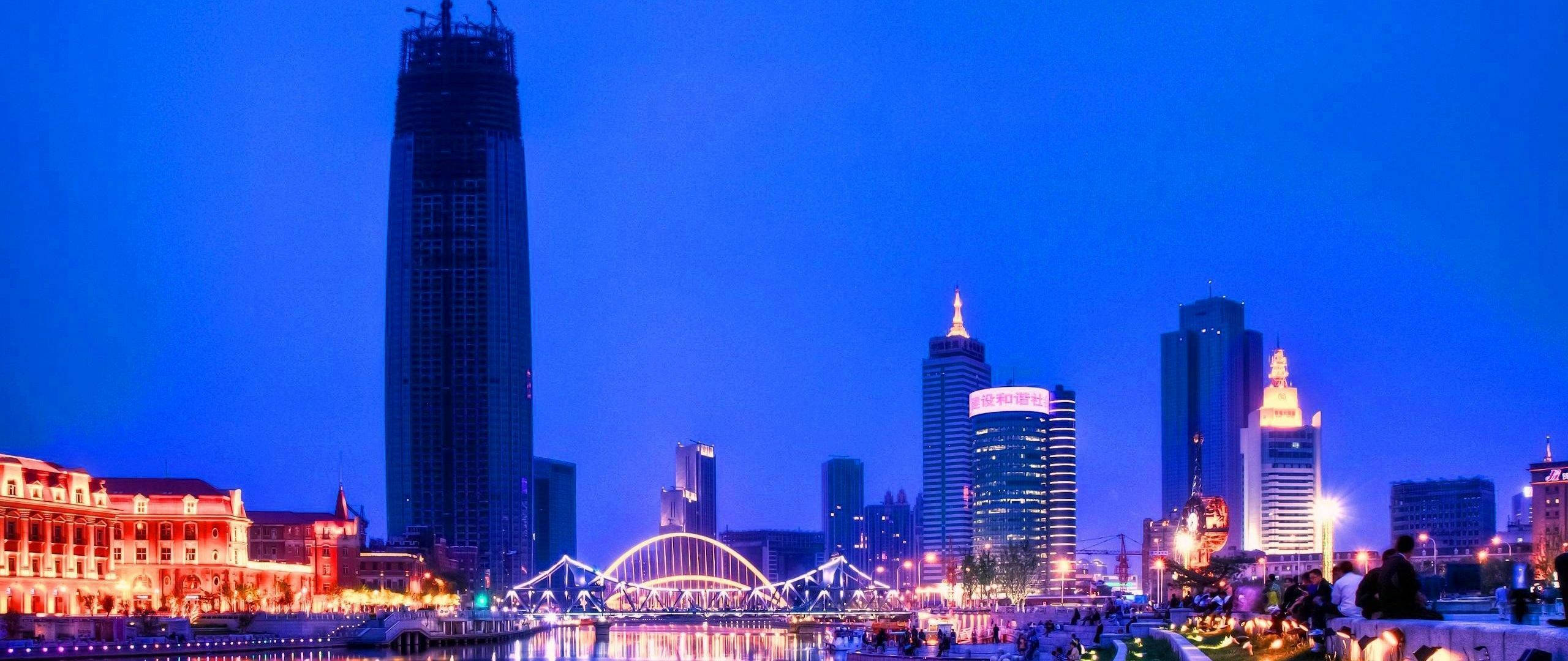 Tianjin 桌布 壁紙 — 黃昏時分充滿活力的城市天際線，燈火通明的摩天大樓和發光的拱橋倒映在平靜的水面上。