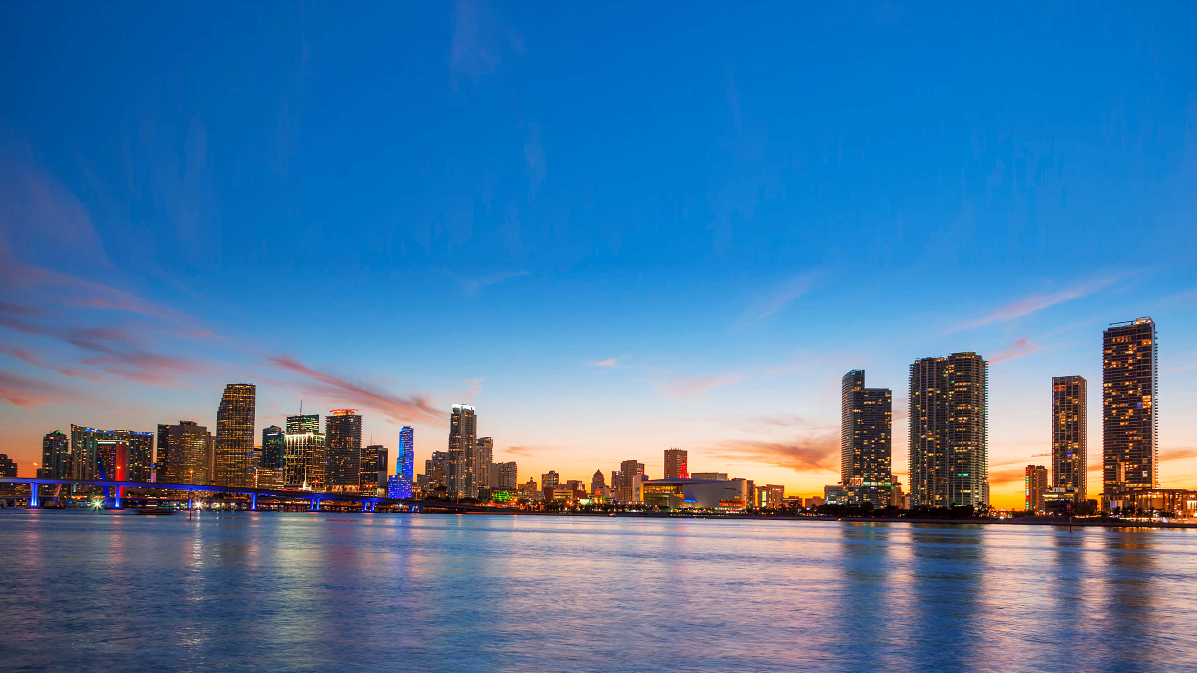 Miami 桌布 壁紙 — 黃昏時分充滿活力的城市天際線，發光的摩天大樓倒映在平靜的水面上，天空呈現藍橙漸變色。