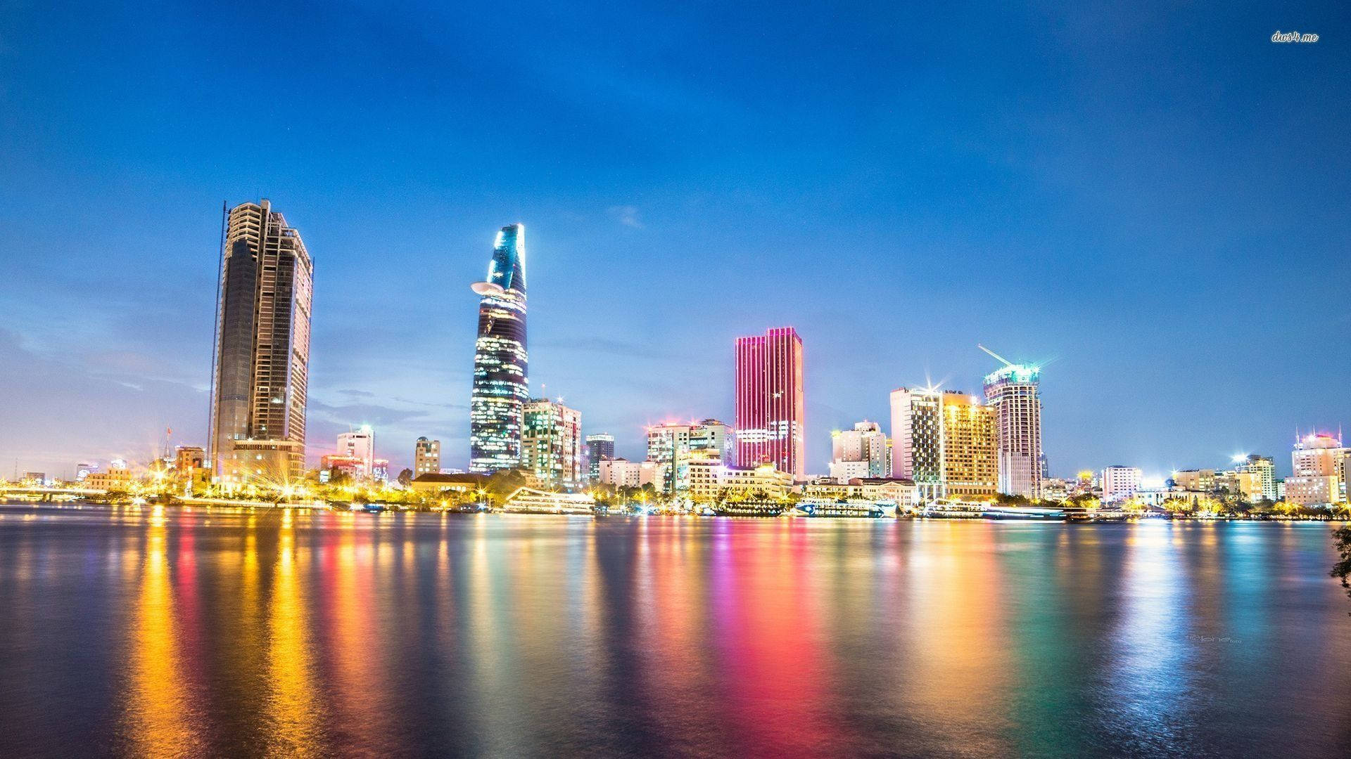 Ho Chi Minh City 桌布 壁紙 — 黃昏時分充滿活力的城市天際線，色彩繽紛的摩天大樓在深藍色天空下平靜的水面上倒映。